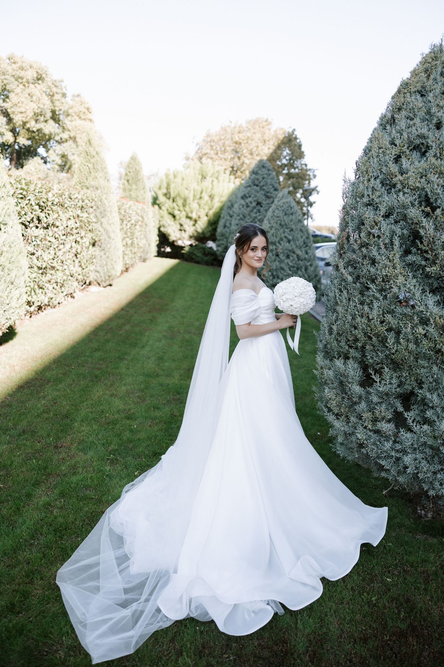 Bride in flowing white gown poses with bouquet along manicured garden path.