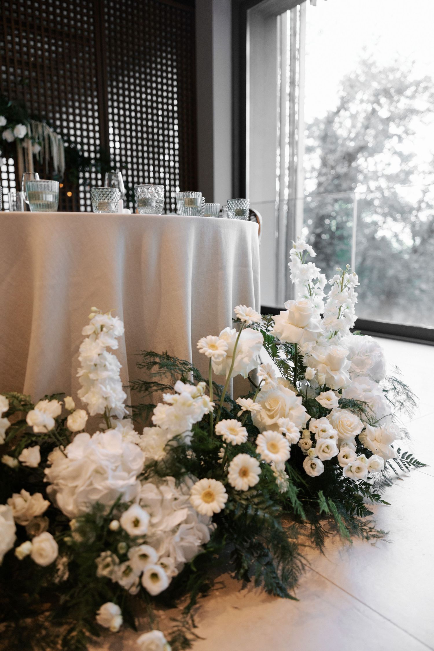 Elegant white floral arrangement with roses and greenery decorating a round table by a window.