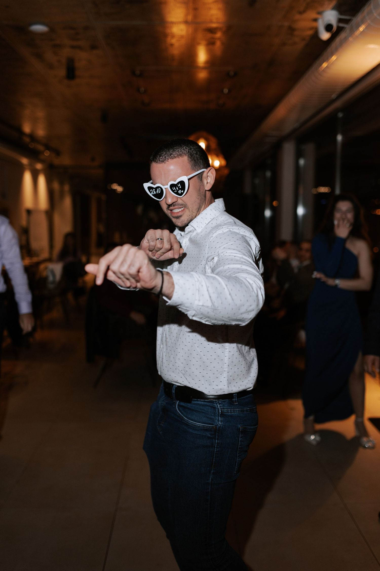 A wedding guest dances enthusiastically wearing heart-shaped sunglasses and a white button-up shirt.