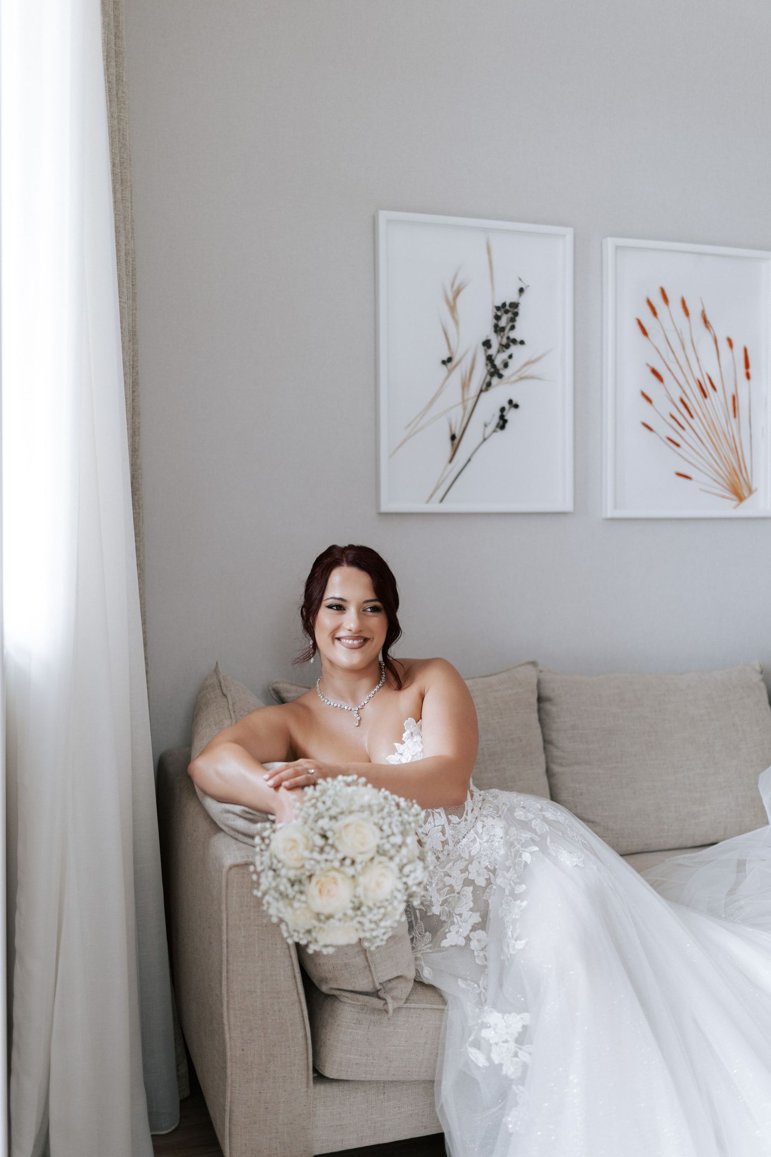 Person in white dress sitting on grey couch holding bouquet with minimal wall art.
