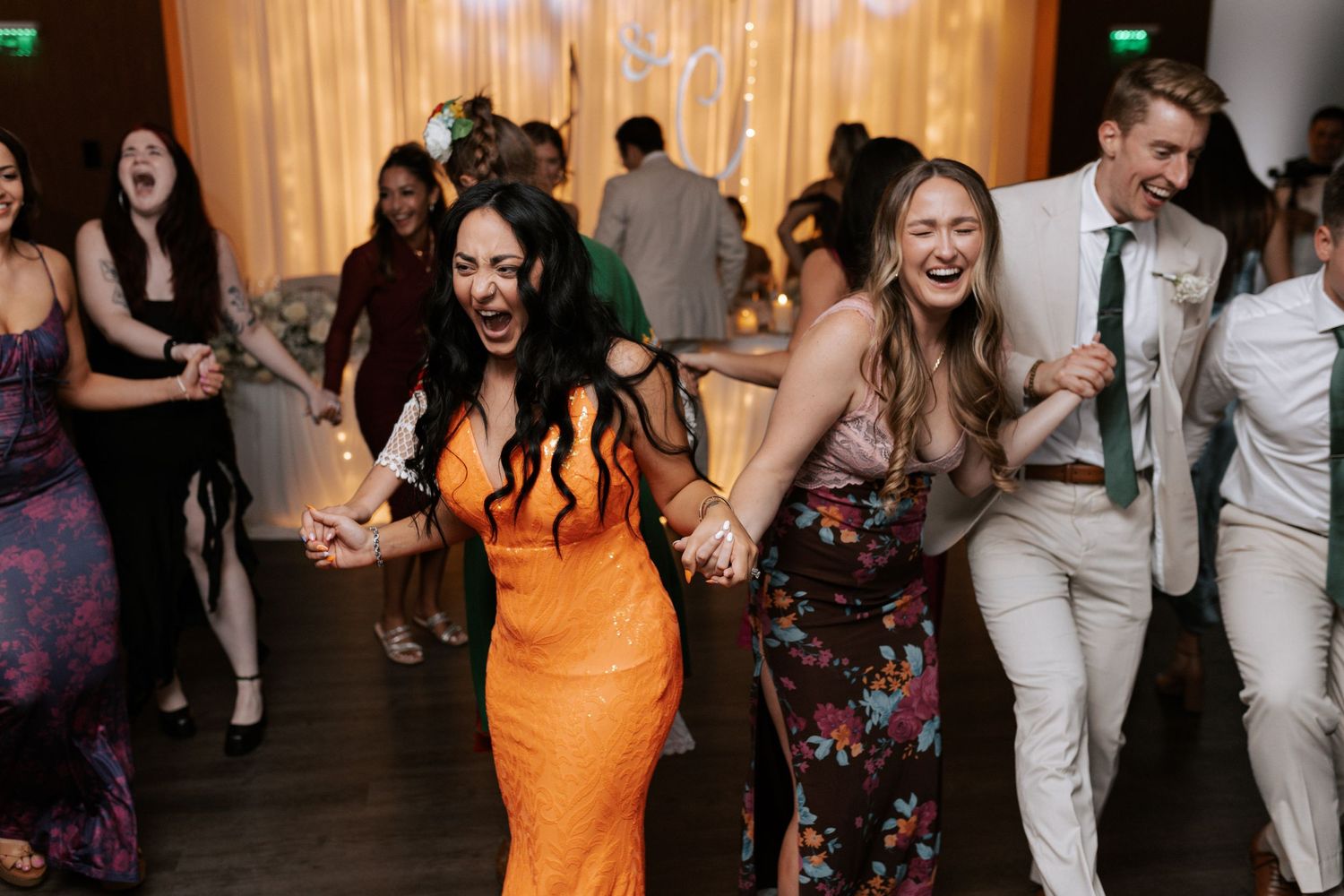 Wedding guests dance joyfully on a wooden dance floor while wearing colorful dresses and formal attire.
