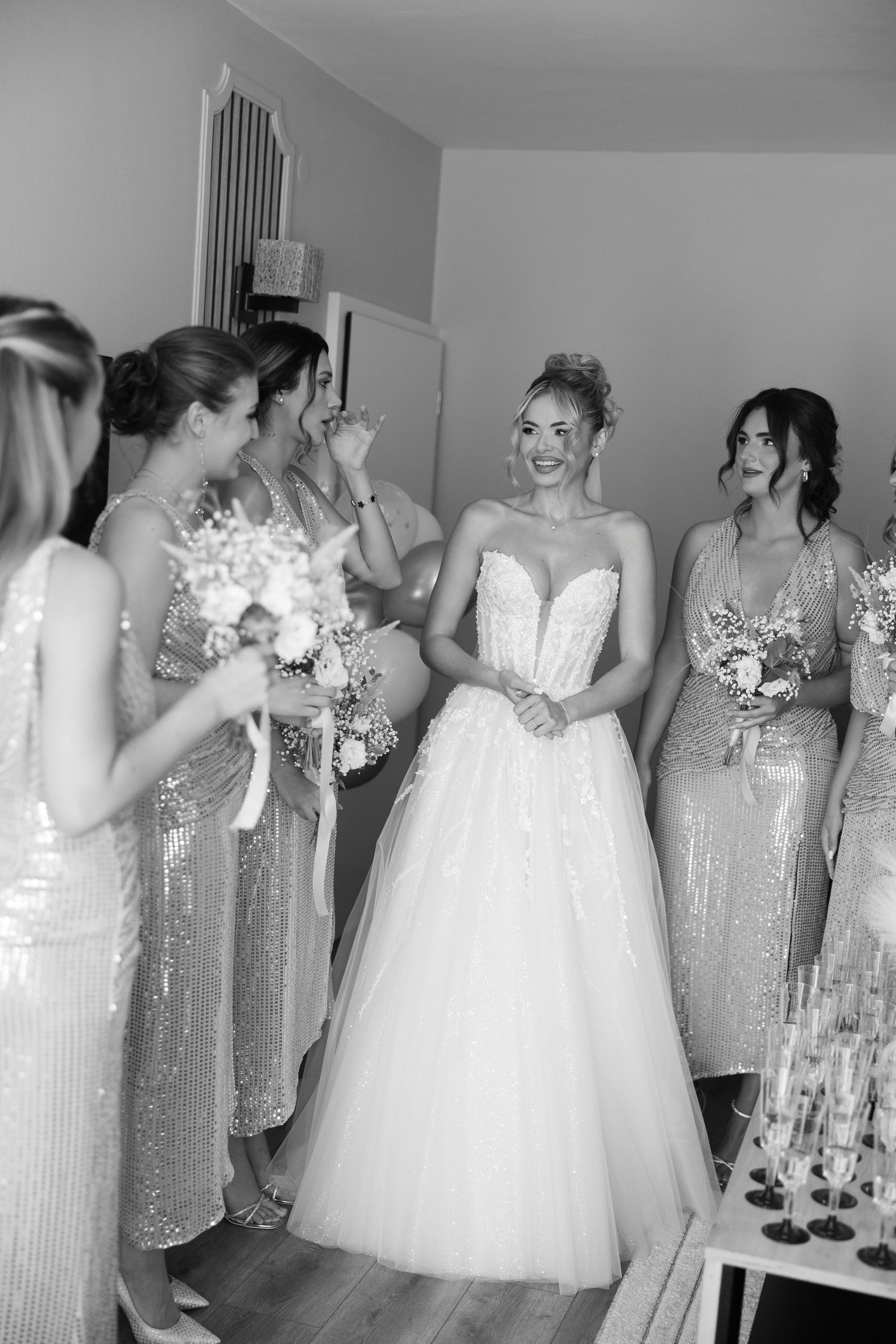 A bride in a white strapless ballgown shares a joyful moment with bridesmaids wearing sparkly champagne dresses.