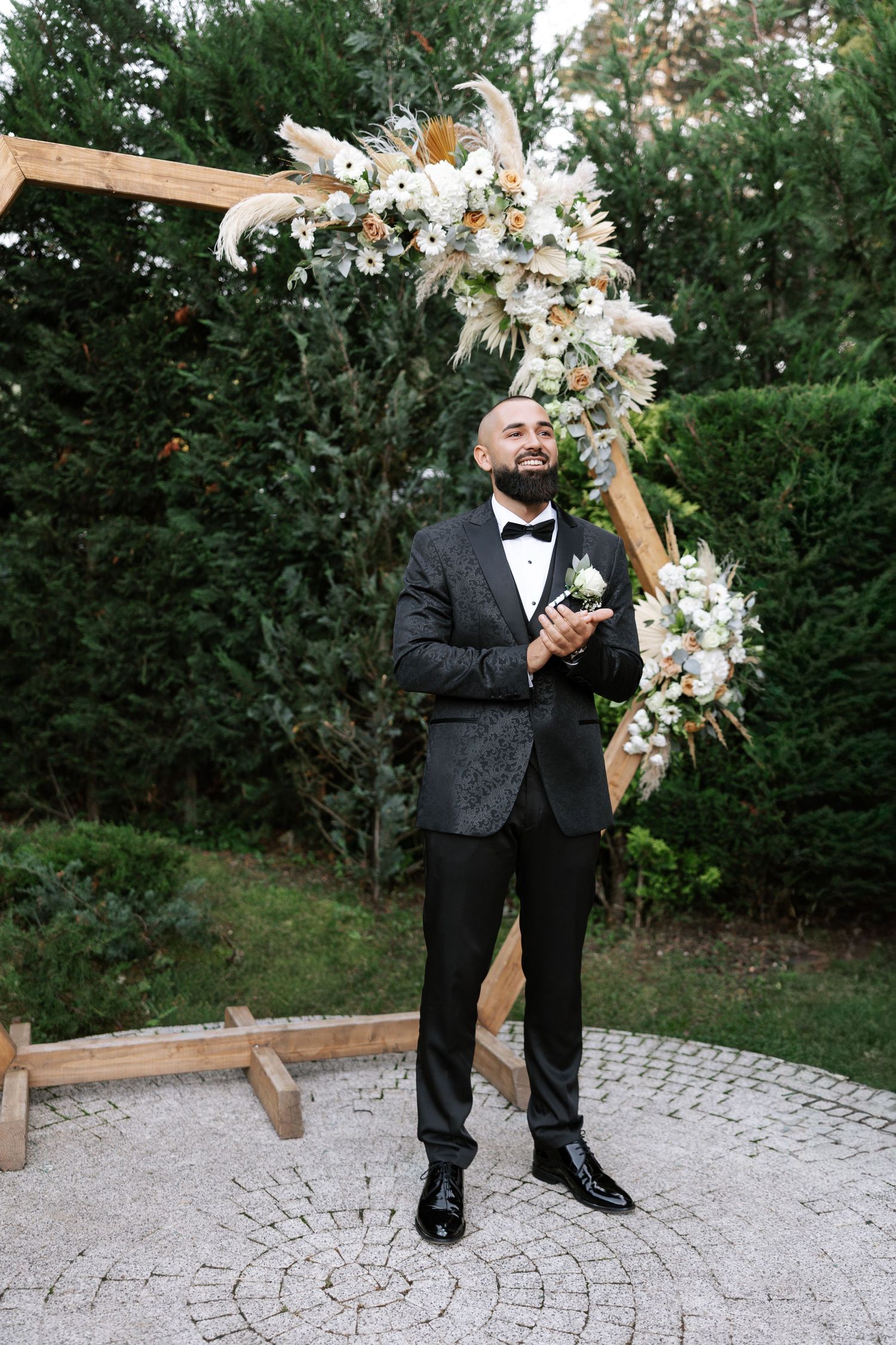 A person in a black tuxedo stands on a stone paver patio next to a wooden arch decorated with white flowers.