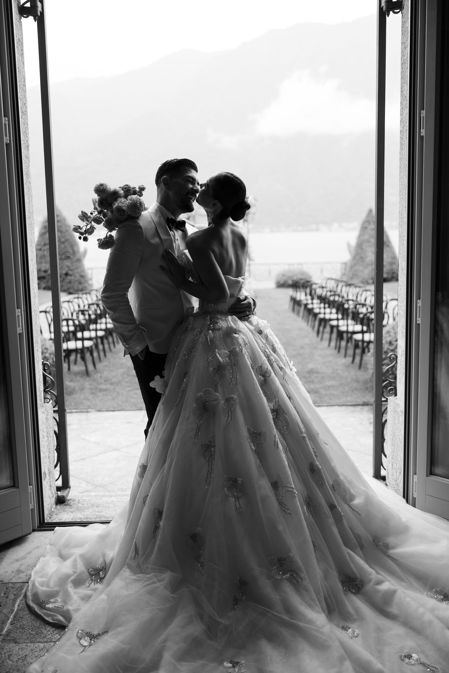 A romantic black and white wedding photo showing a couple in an elegant doorway overlooking rows of chairs outdoors.