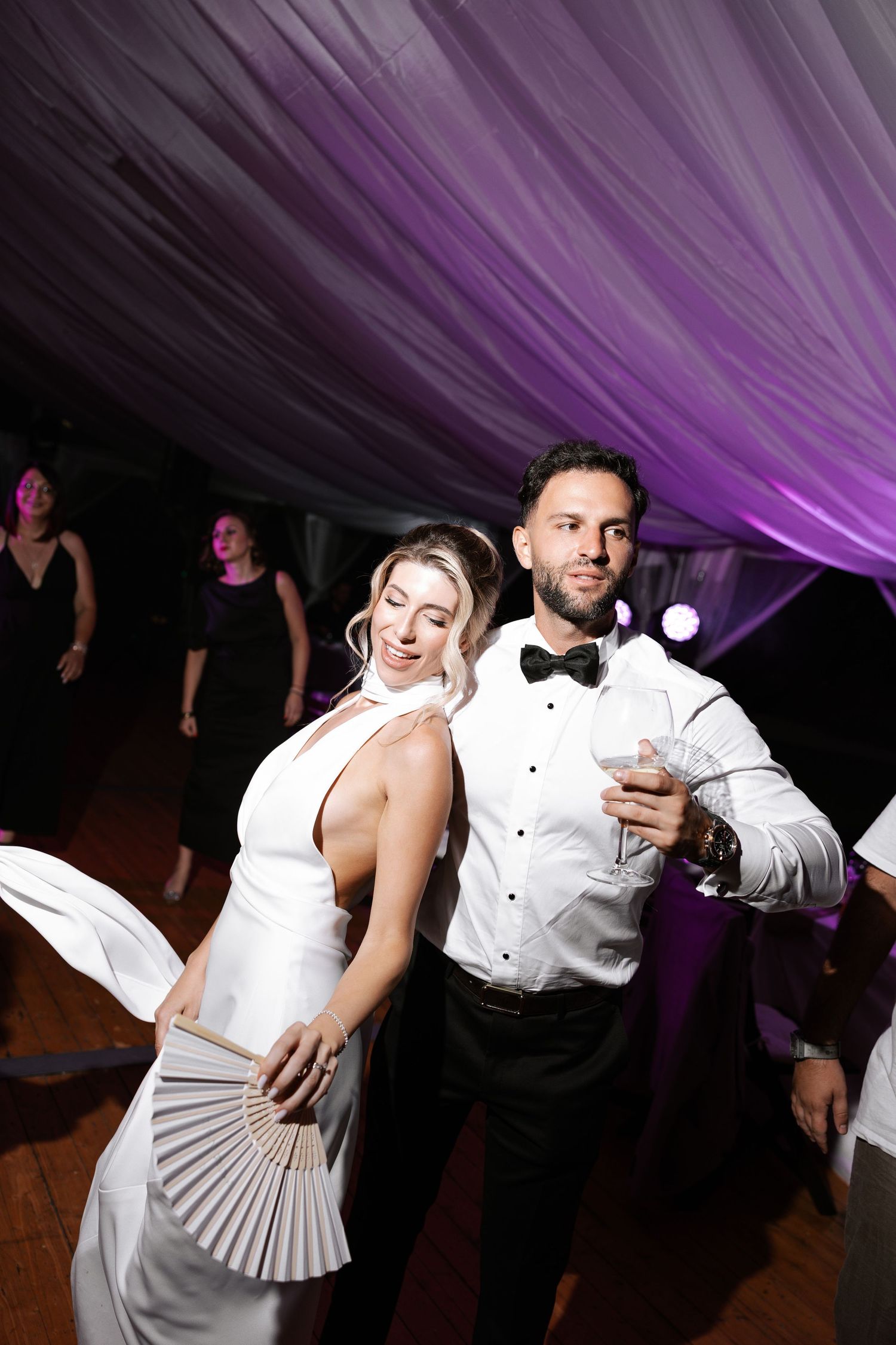 Wedding party guests dance energetically on the dance floor under dramatic purple lighting at a reception celebration.