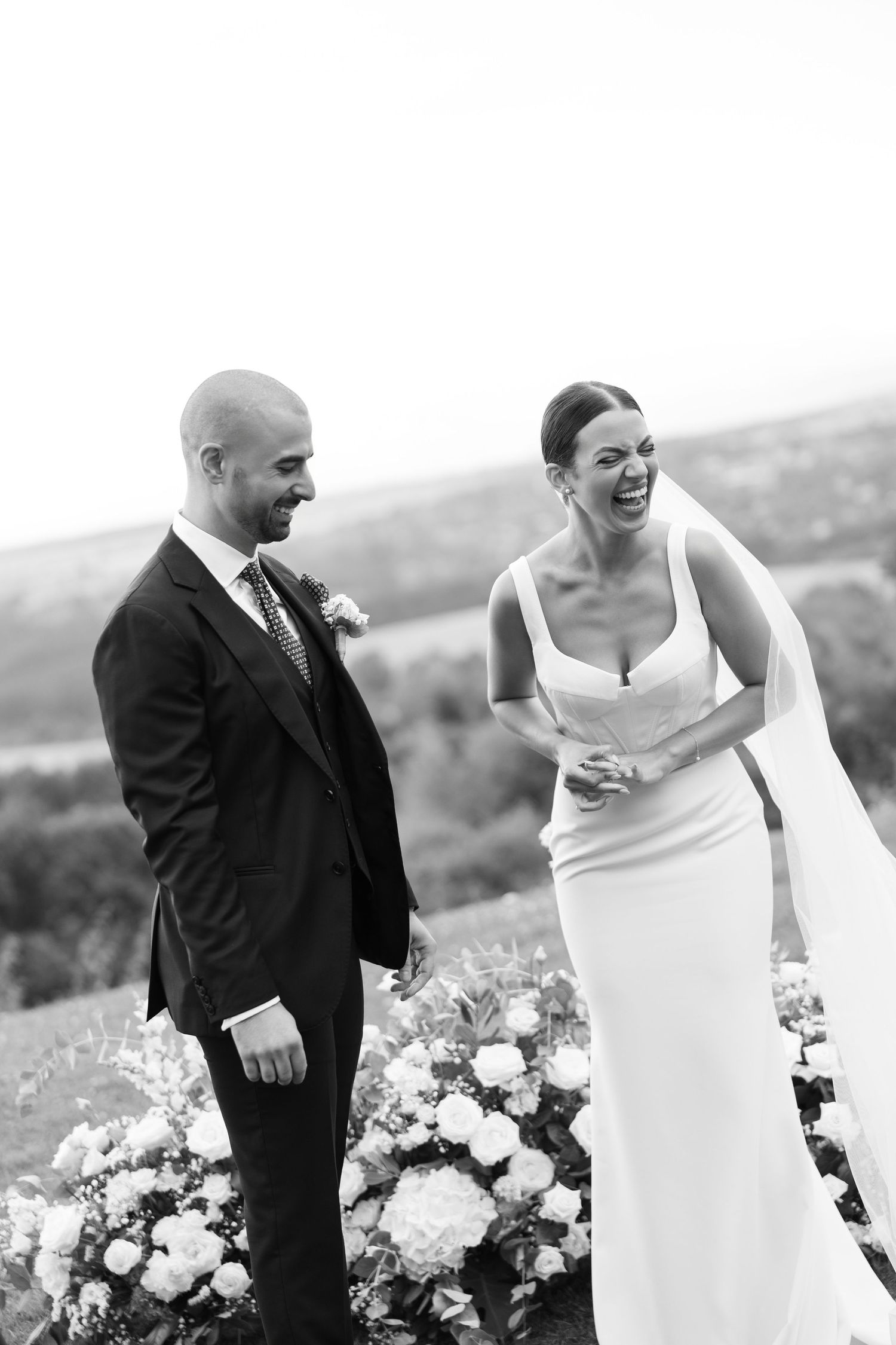 A bride and groom share joyful laughter during their outdoor wedding ceremony with floral arrangements in the background.