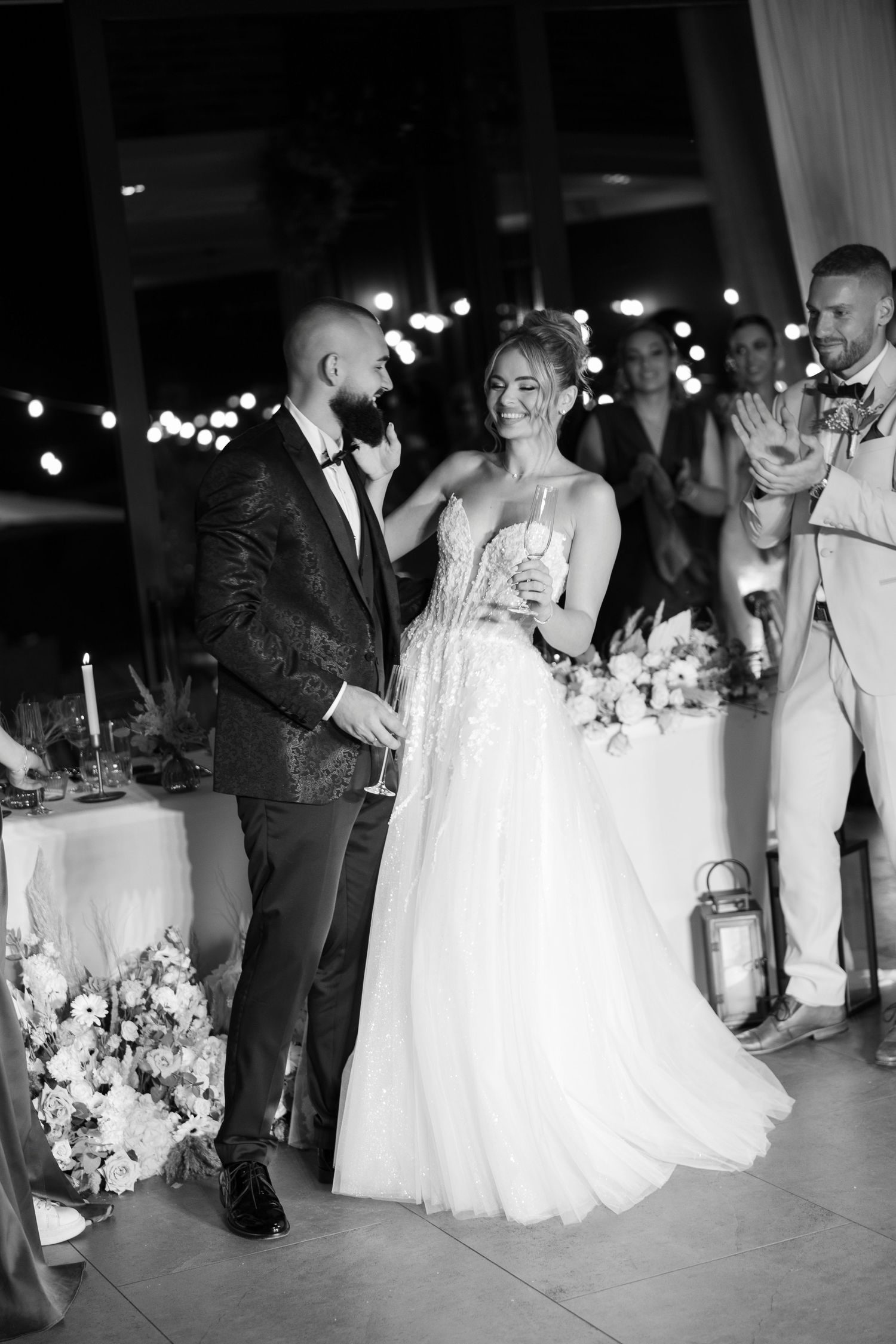 Black and white wedding photo of a couple sharing a joyful moment during their reception with soft lighting and floral decor.