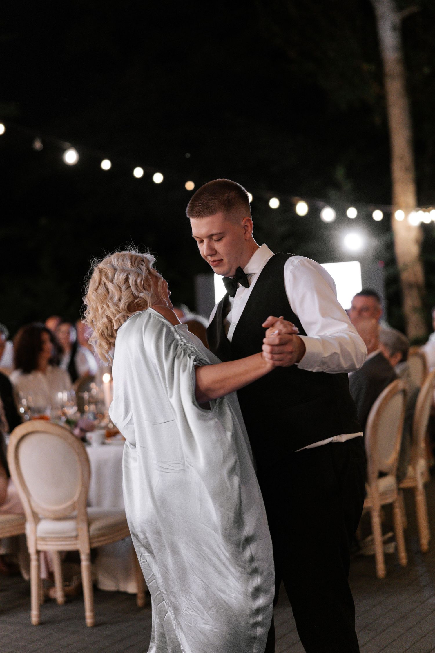 Wedding guests dancing in formal attire under string lights at an outdoor reception.