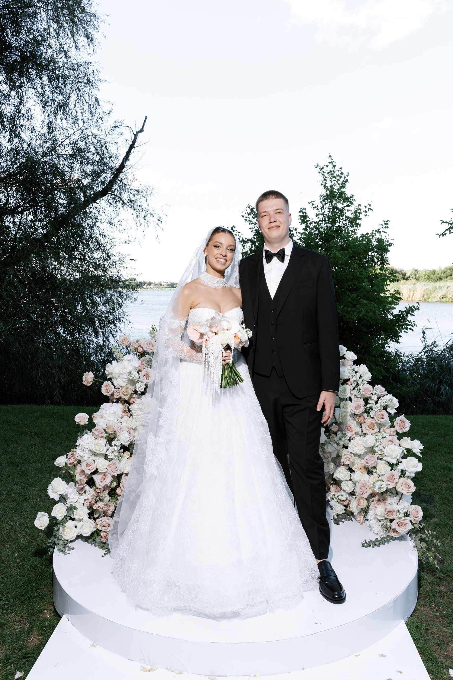 Newlyweds pose under a floral arch with pink and white roses against a waterfront backdrop.