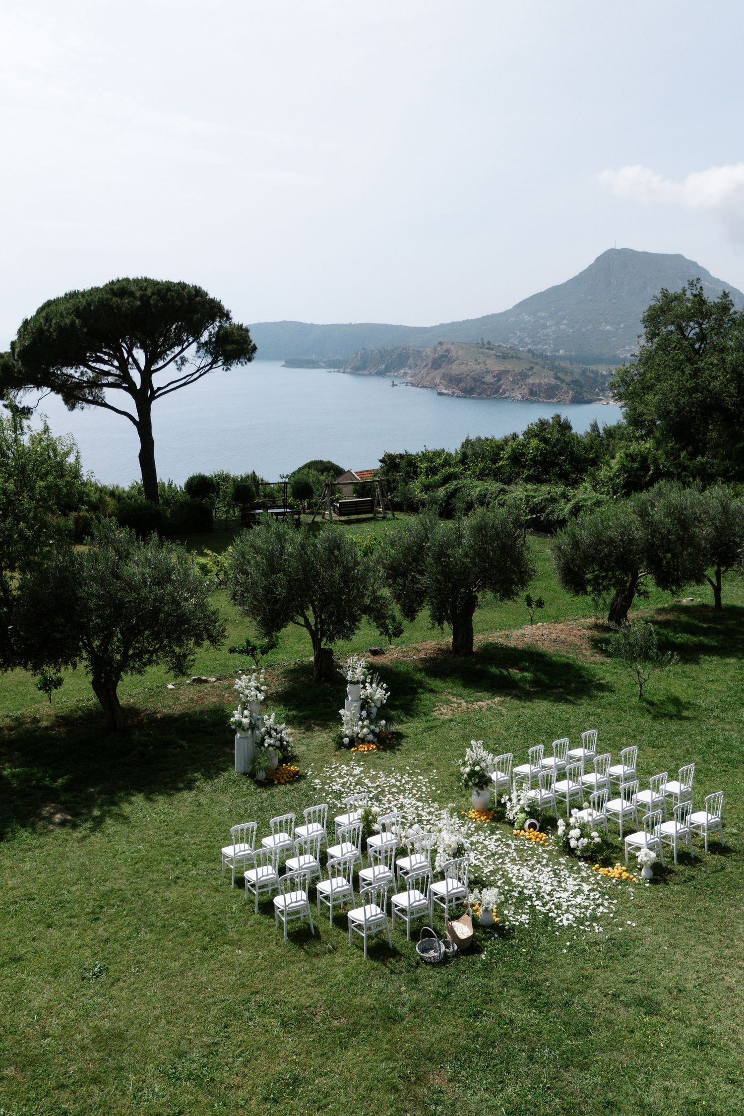 White chairs arranged for an outdoor wedding ceremony on a grassy hillside overlooking coastal mountains and sea views.