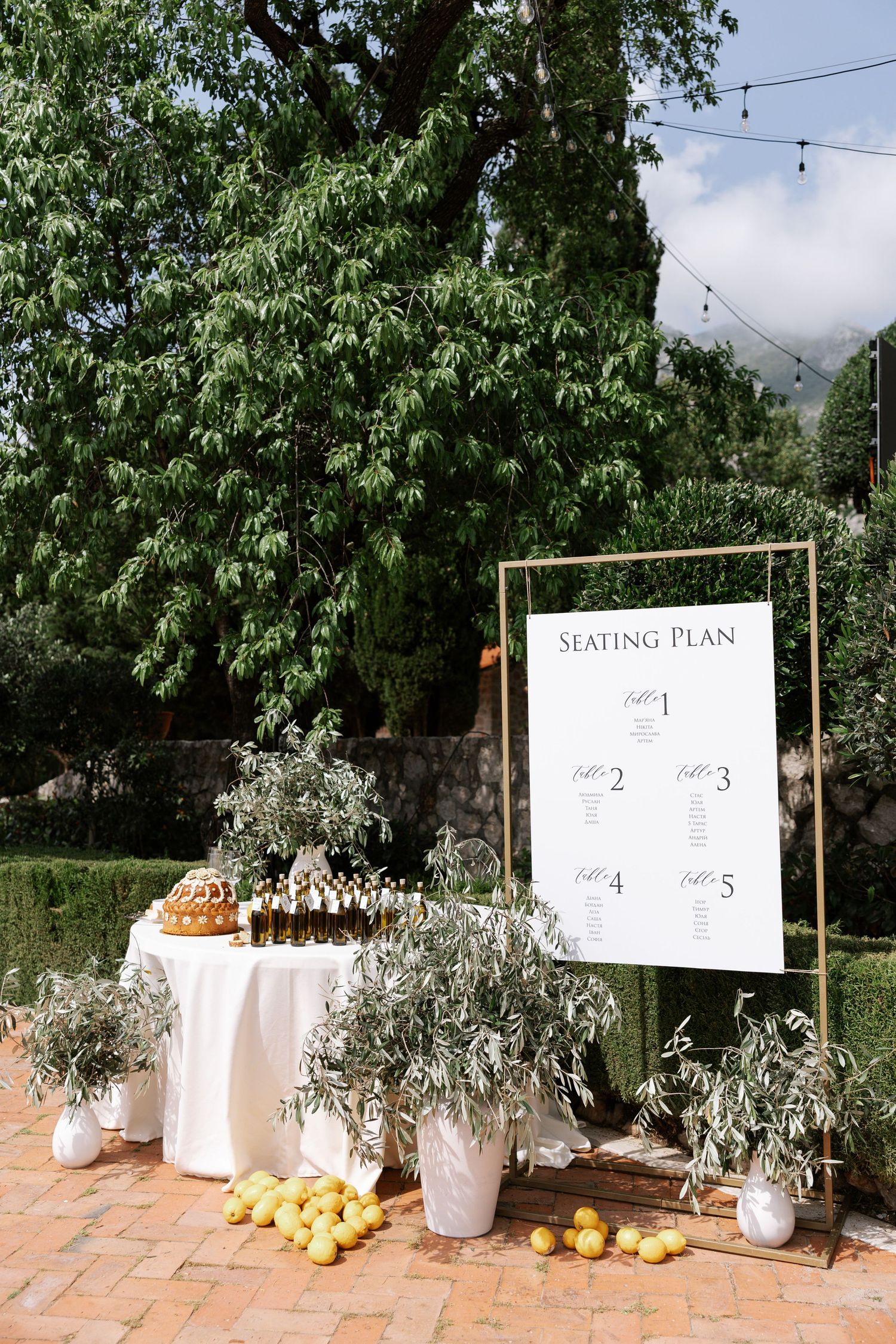 Rustic outdoor wedding seating chart display with olive branches and citrus fruits on terracotta tiles.