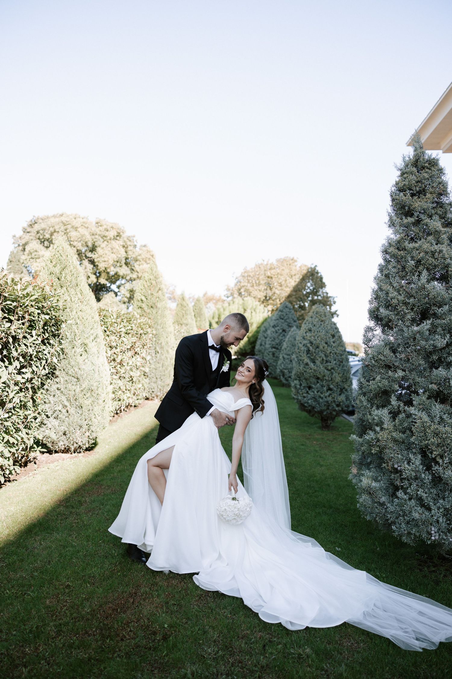 Dramatic garden wedding photo with couple embracing among evergreen trees.