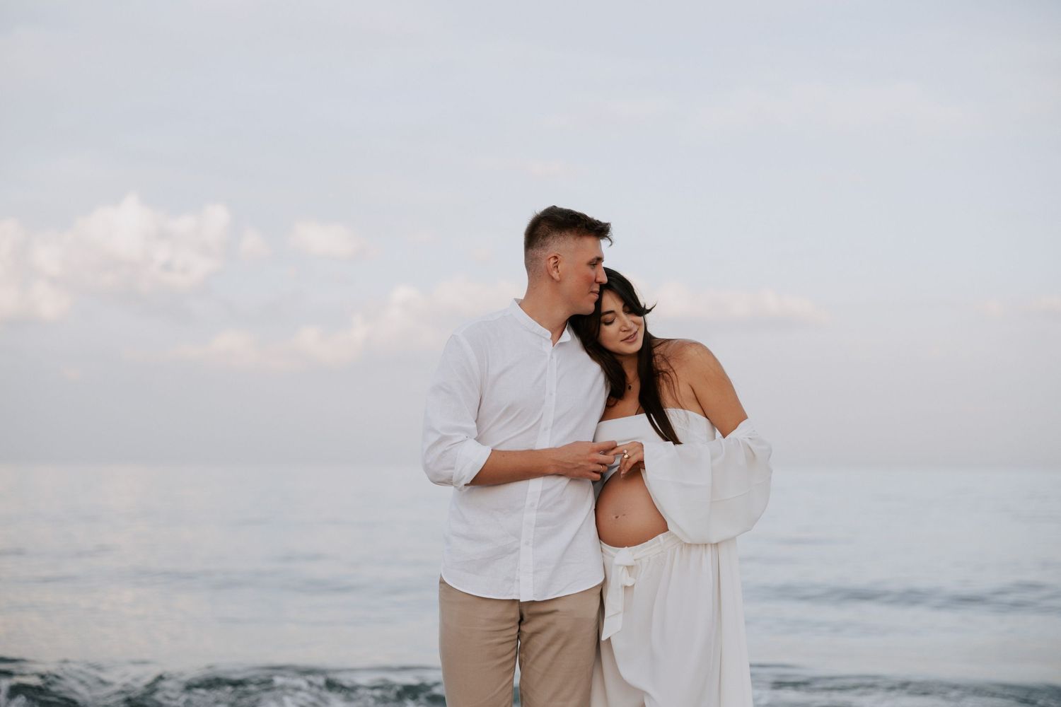 An intimate maternity beach photo of a couple in white clothing against the ocean.
