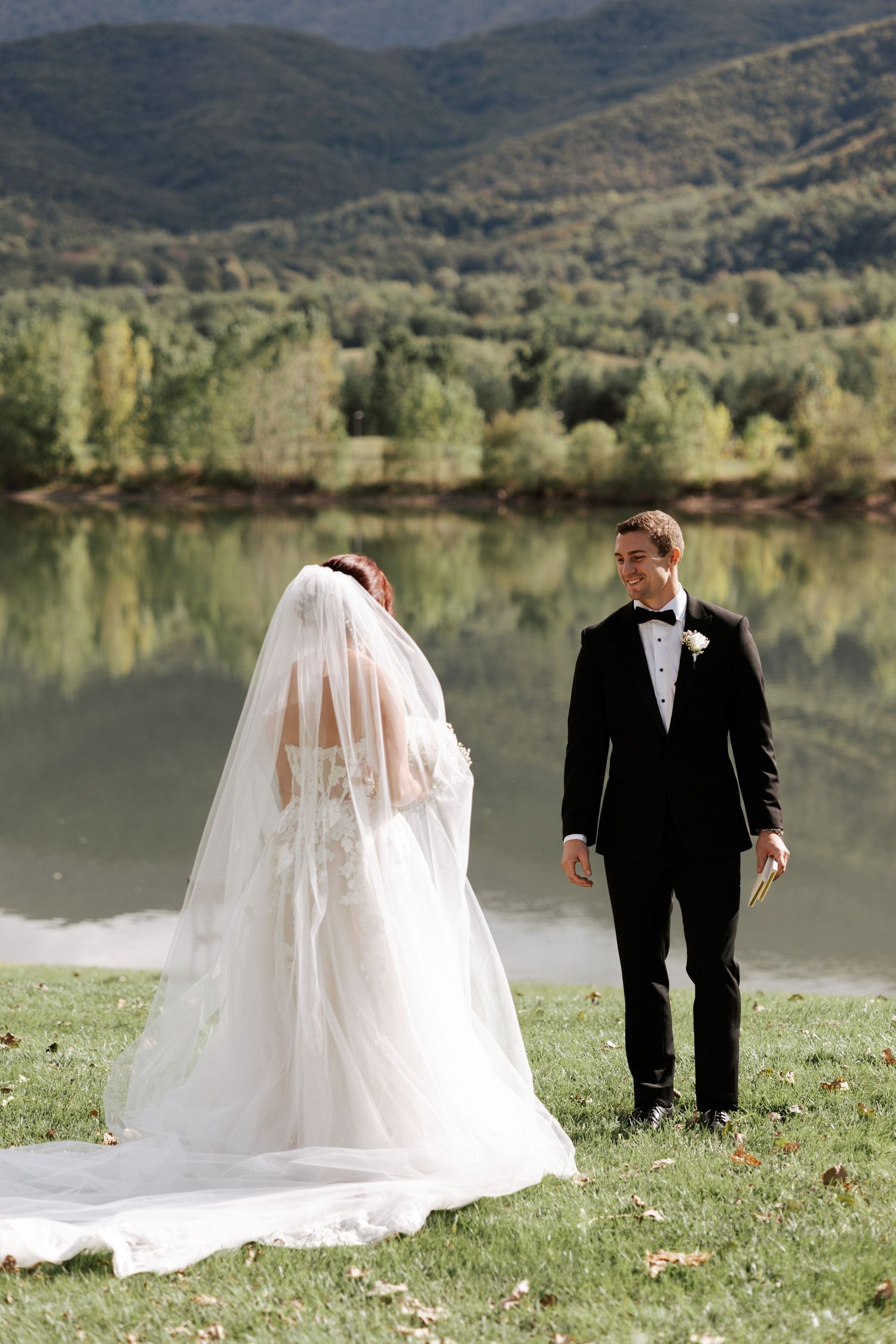 A bride and groom stand on a grassy hillside overlooking a tranquil mountain lake.