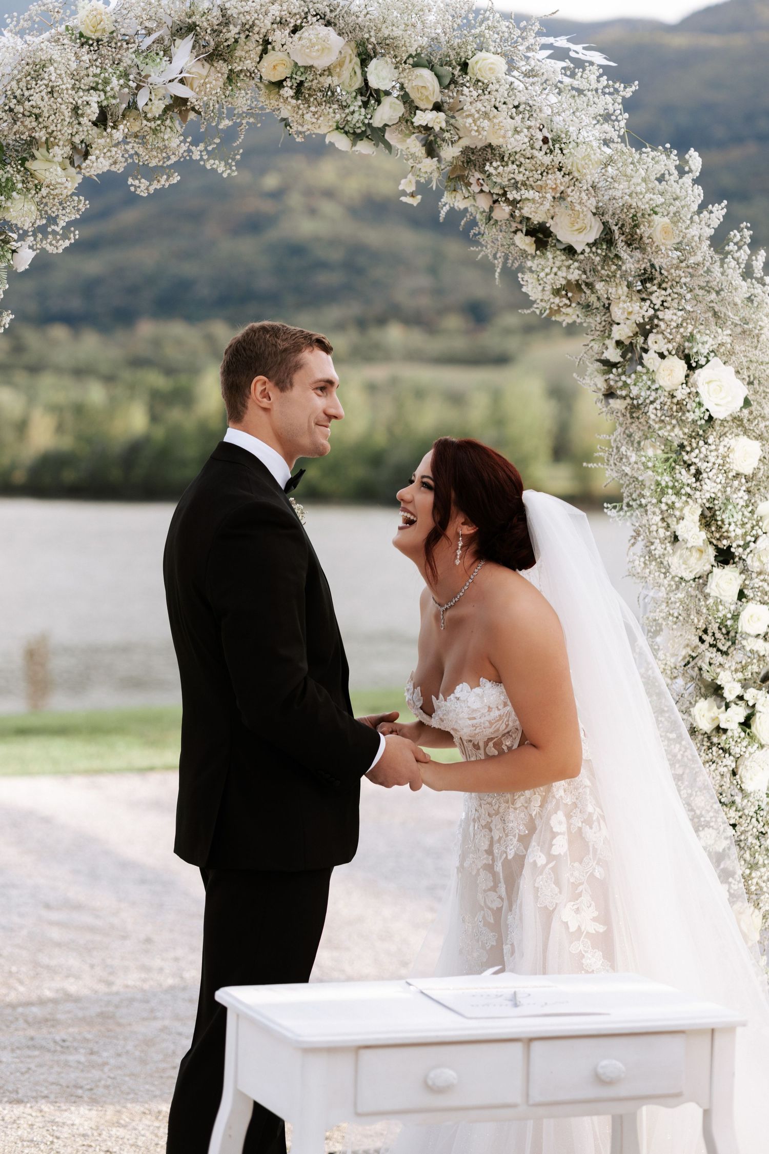 A couple shares a moment under a white floral arch during their mountain wedding ceremony.