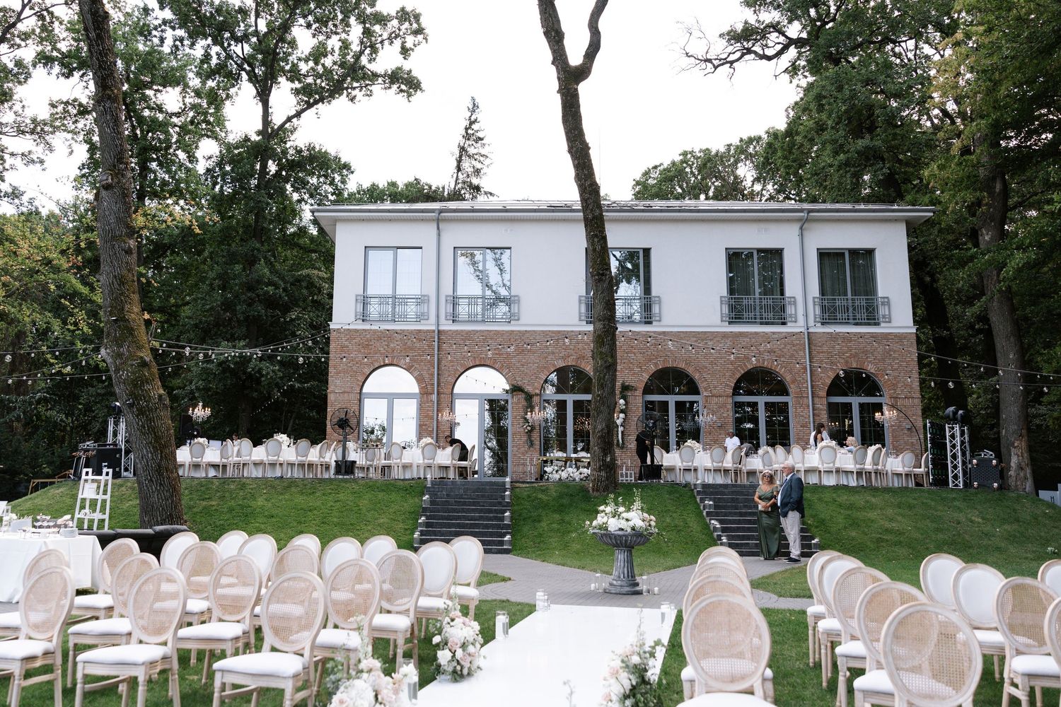 White chairs arranged outdoors for a wedding ceremony with trees and a brick building in the background.