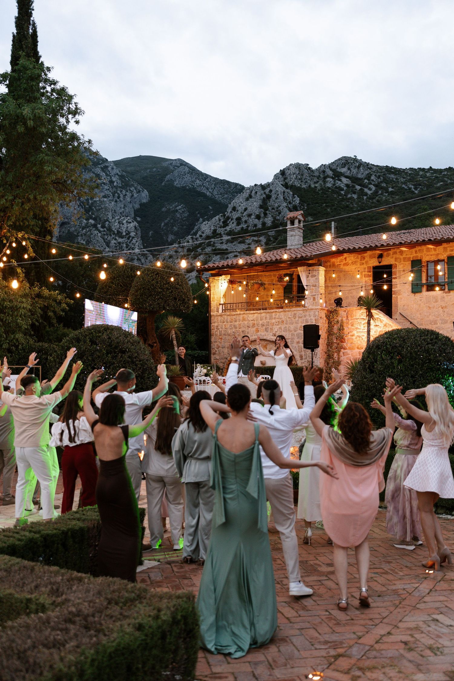 Wedding guests dance together under string lights at an outdoor reception with mountains in the background at dusk.