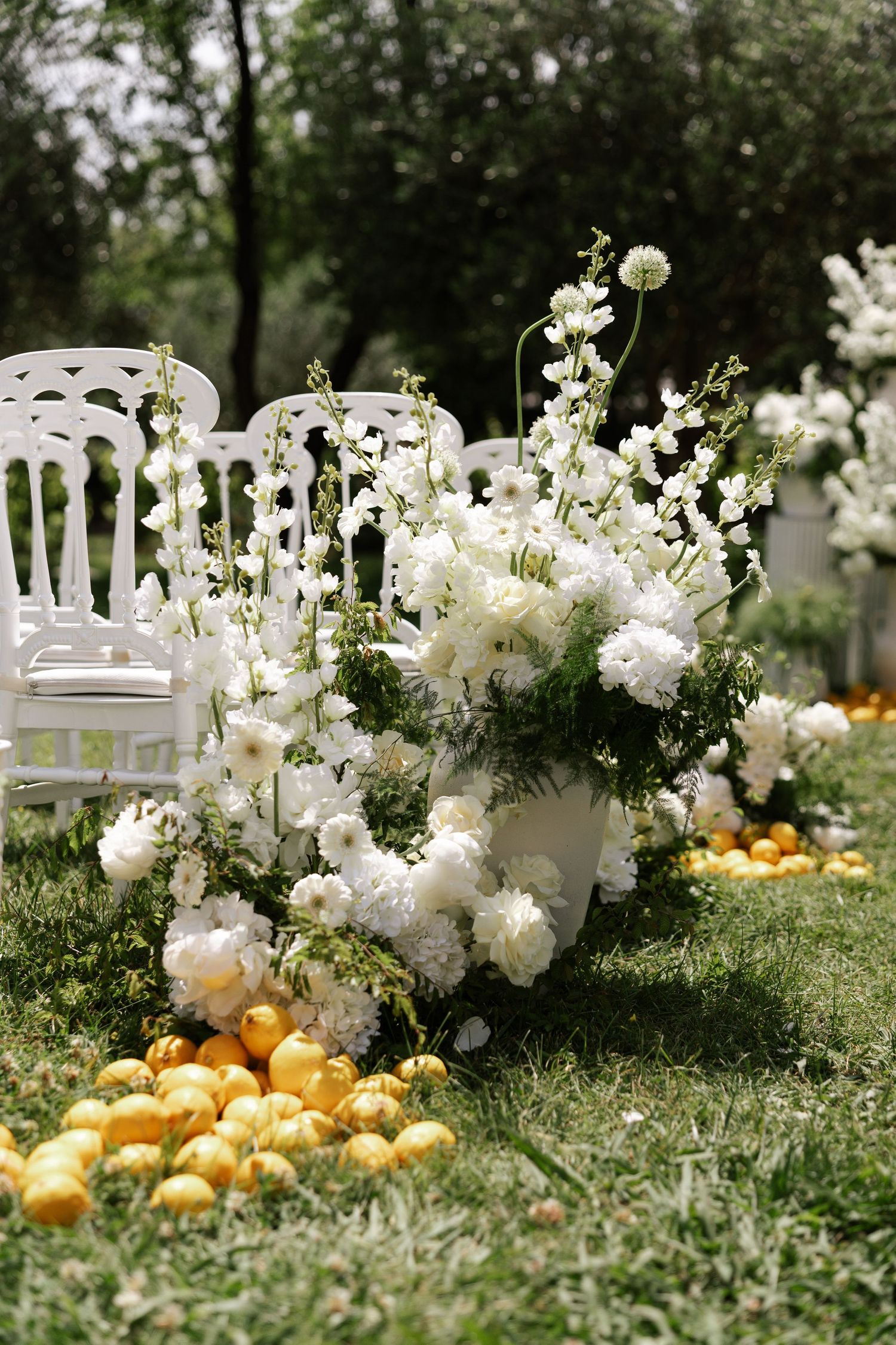White floral arrangements and scattered oranges create an elegant garden party setting with white chairs on lush grass.