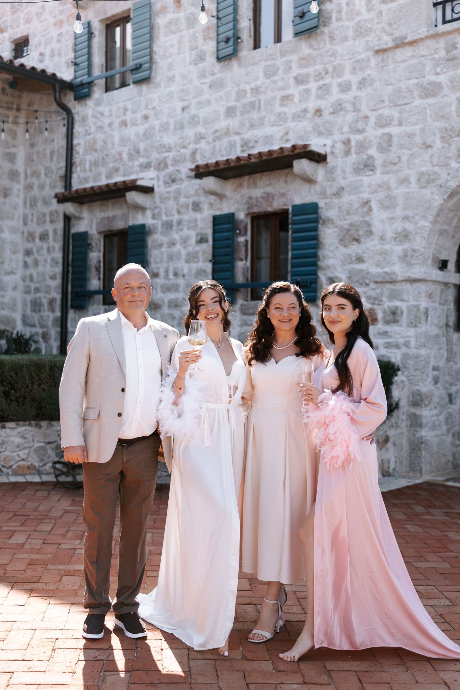A family group poses together outdoors against a stone wall wearing white and pink vintage-style clothing.
