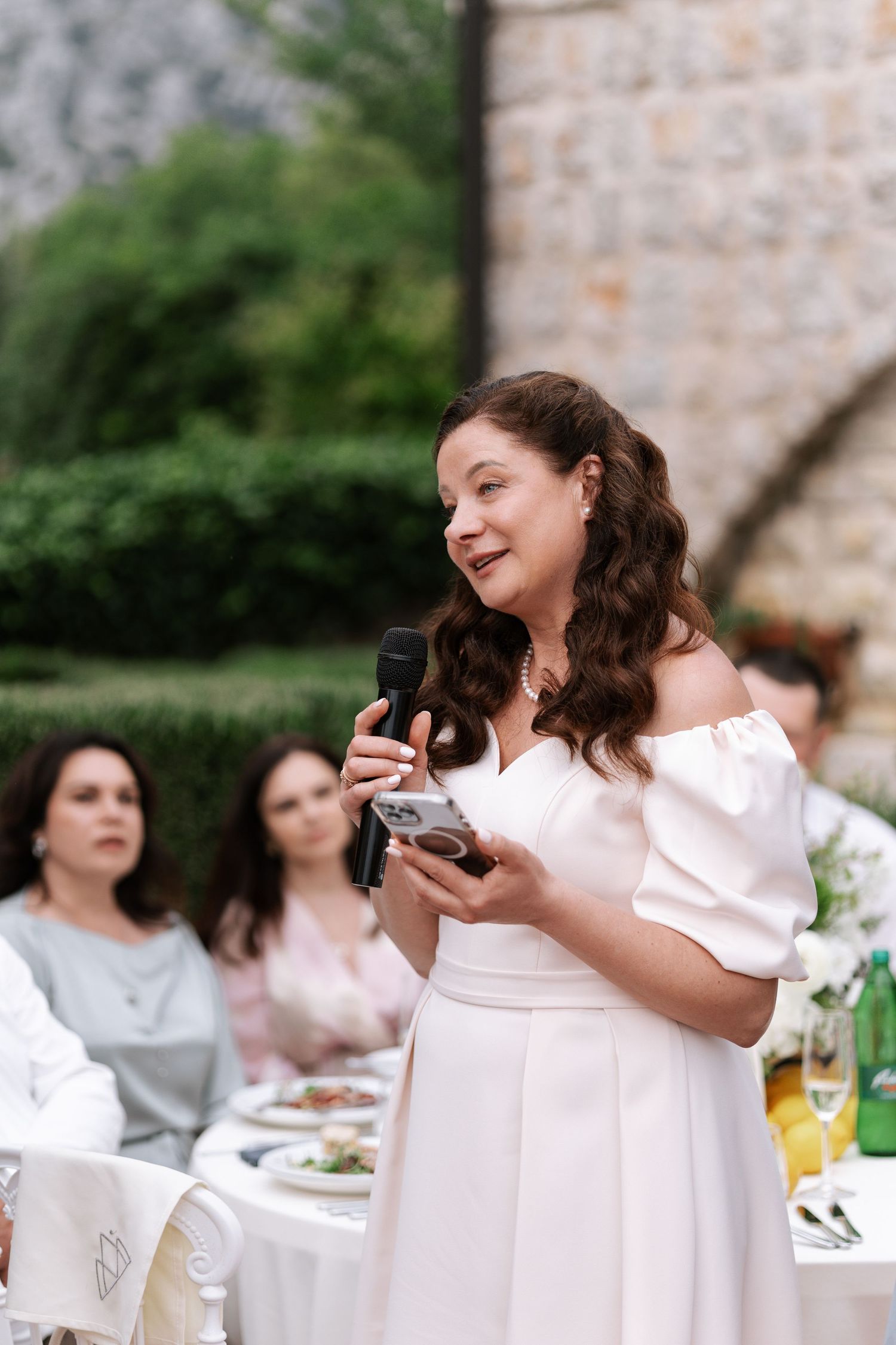 Someone in a white off-shoulder dress gives a speech at an outdoor wedding reception while holding a microphone.