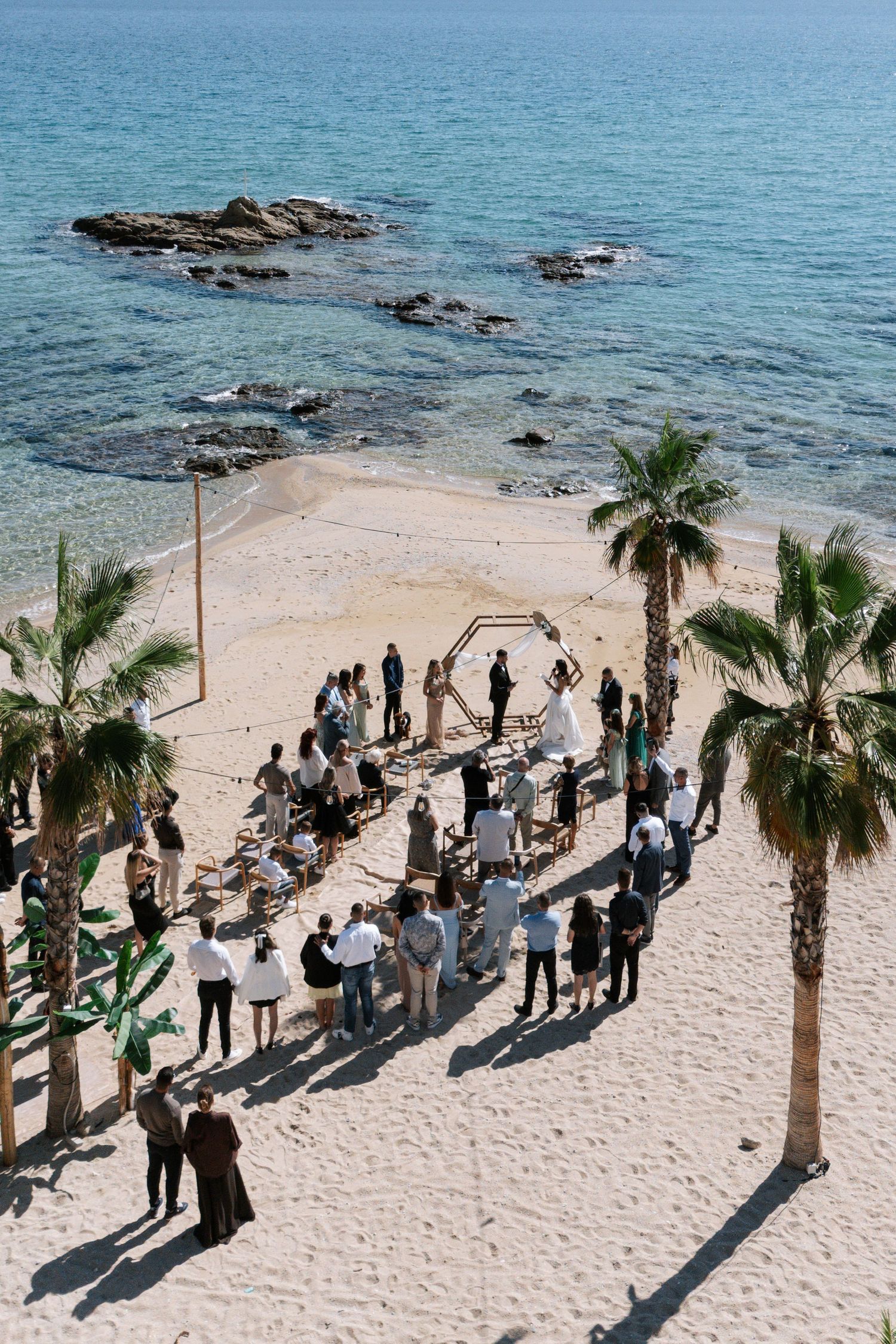 Beachside wedding ceremony setup with palm trees and guests gathered on sandy shore overlooking blue ocean waters.