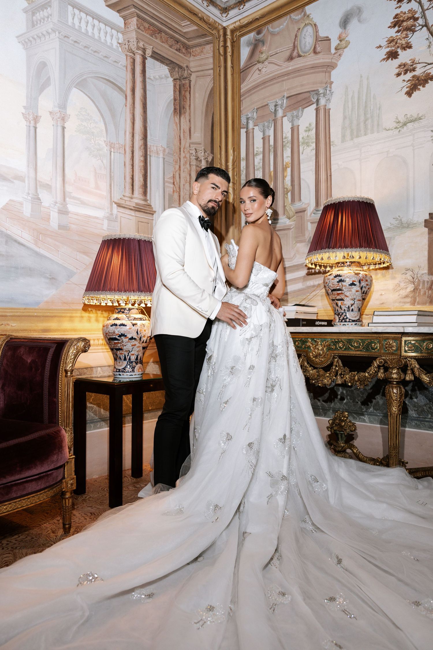 Elegant couple in formal wedding attire pose in luxurious room with ornate decor and vintage table lamps.