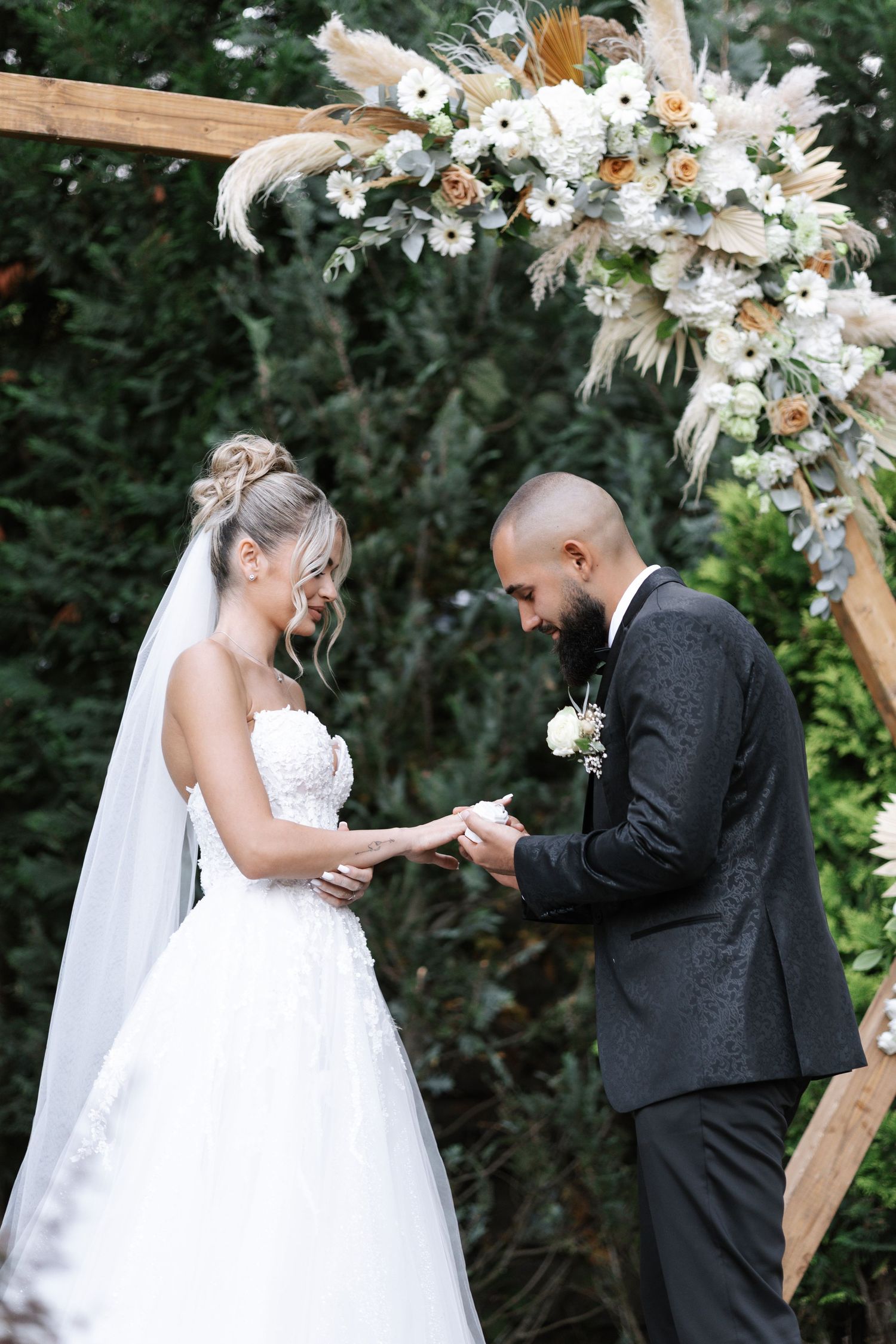 A beautiful outdoor wedding ceremony shows a couple exchanging rings under a floral arch decorated with white flowers.