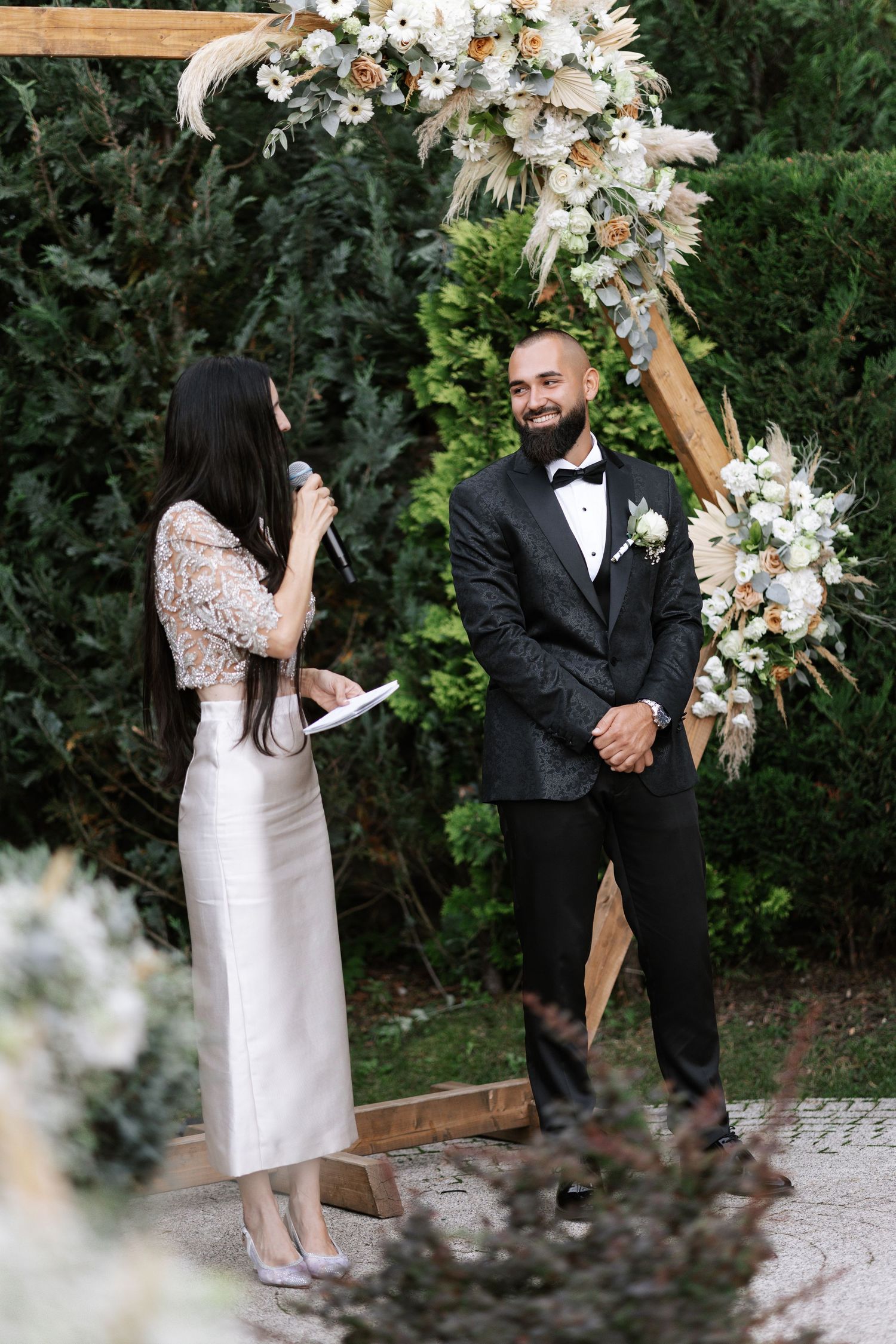 A couple stands before a rustic wooden arch decorated with white flowers and greenery during an outdoor wedding ceremony.