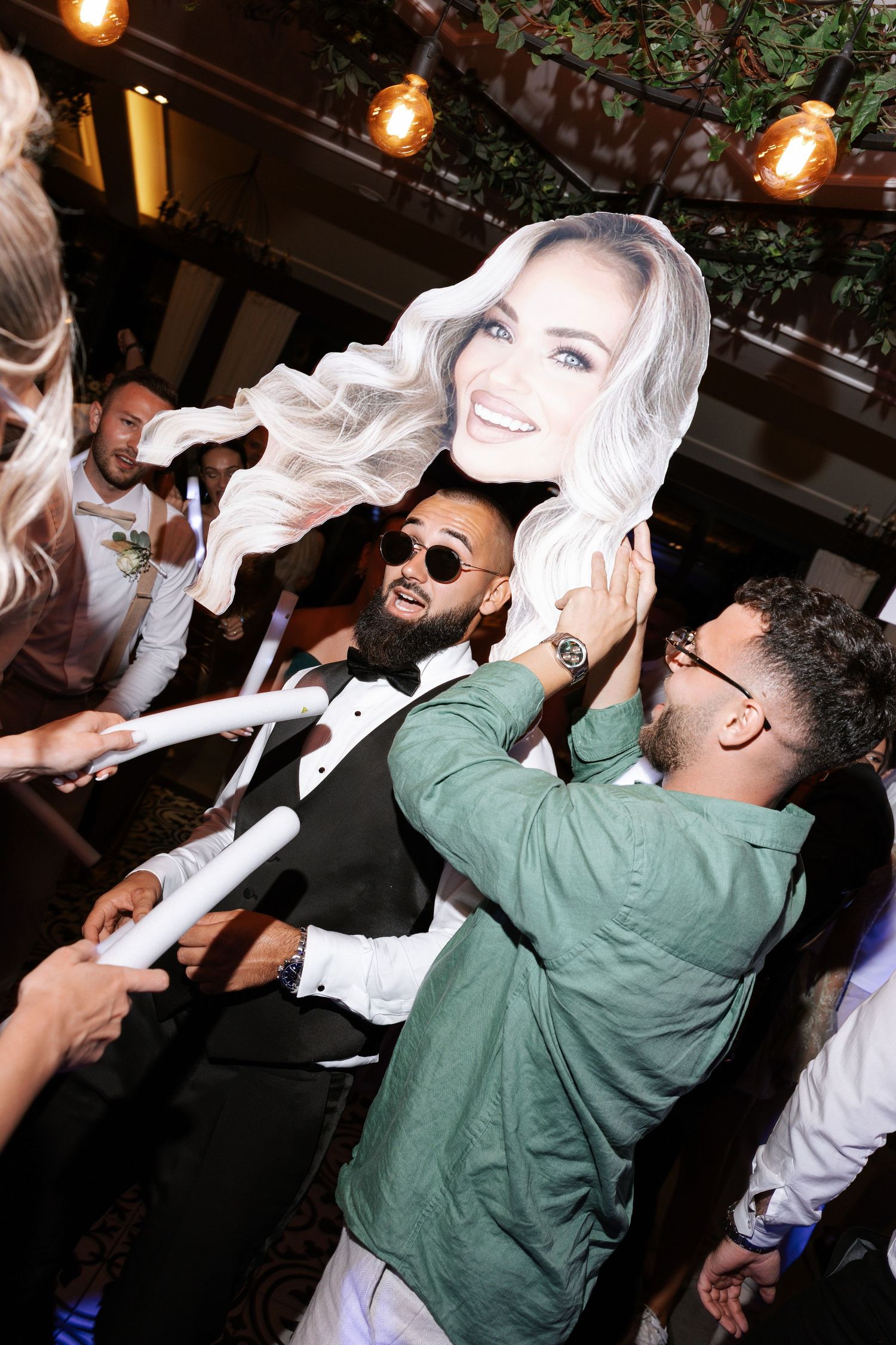 Partygoers at a nightclub hold up and celebrate with a large cardboard cutout head under string lights.