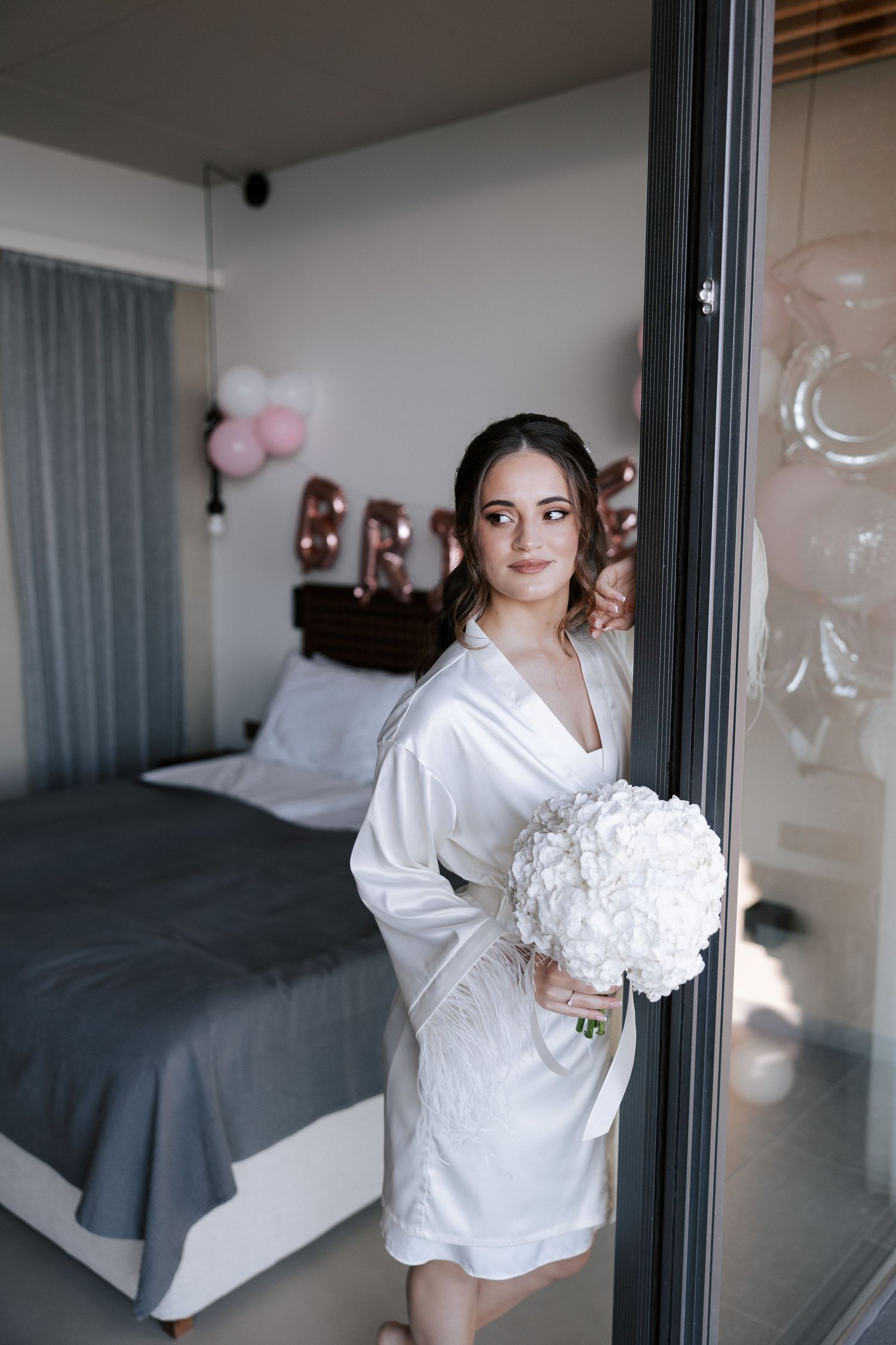 Figure in white robe holding bouquet while posing in modern bedroom setting.