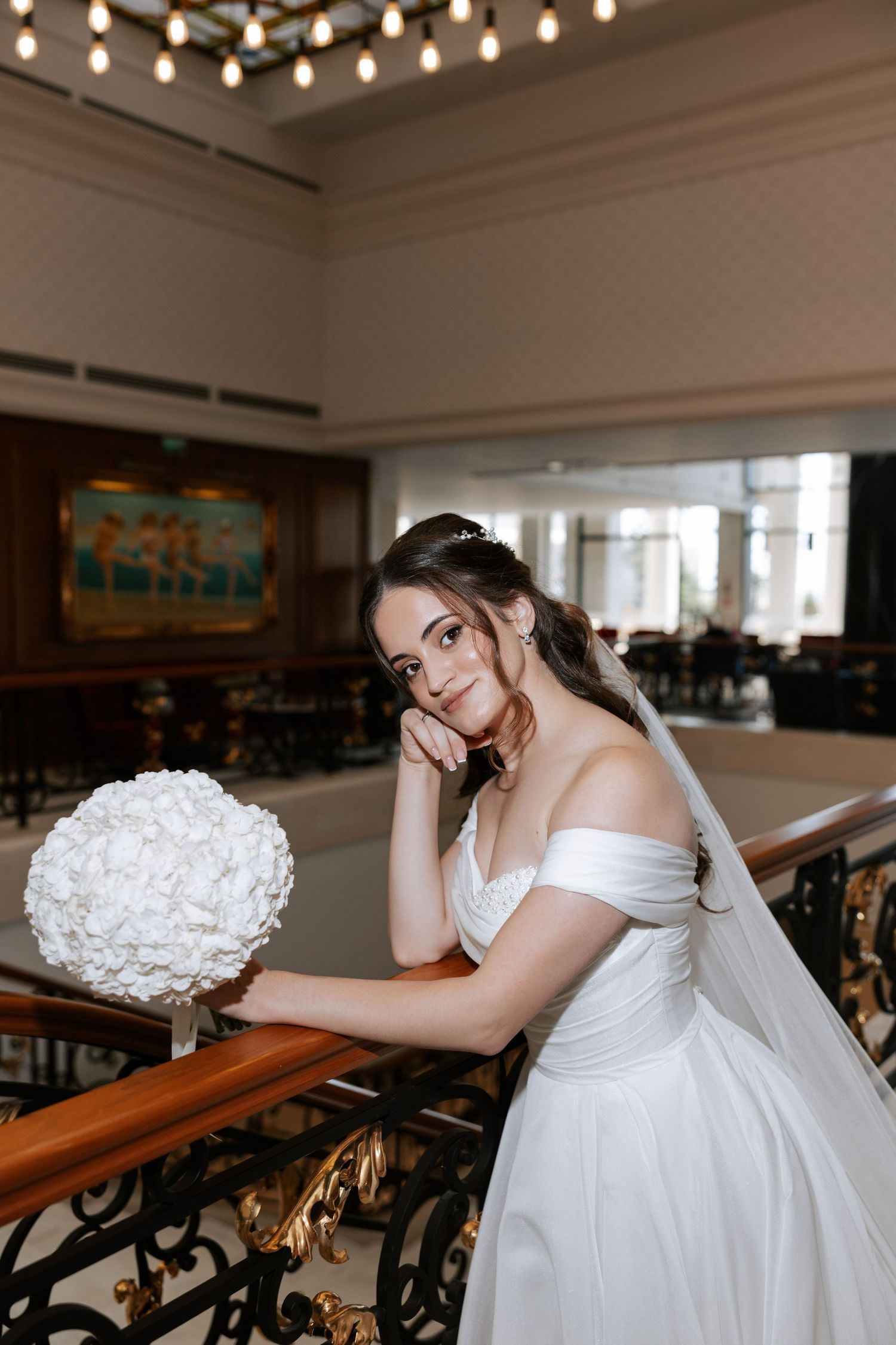 Bride in off-shoulder gown poses with white bouquet by ornate railing under string lights.