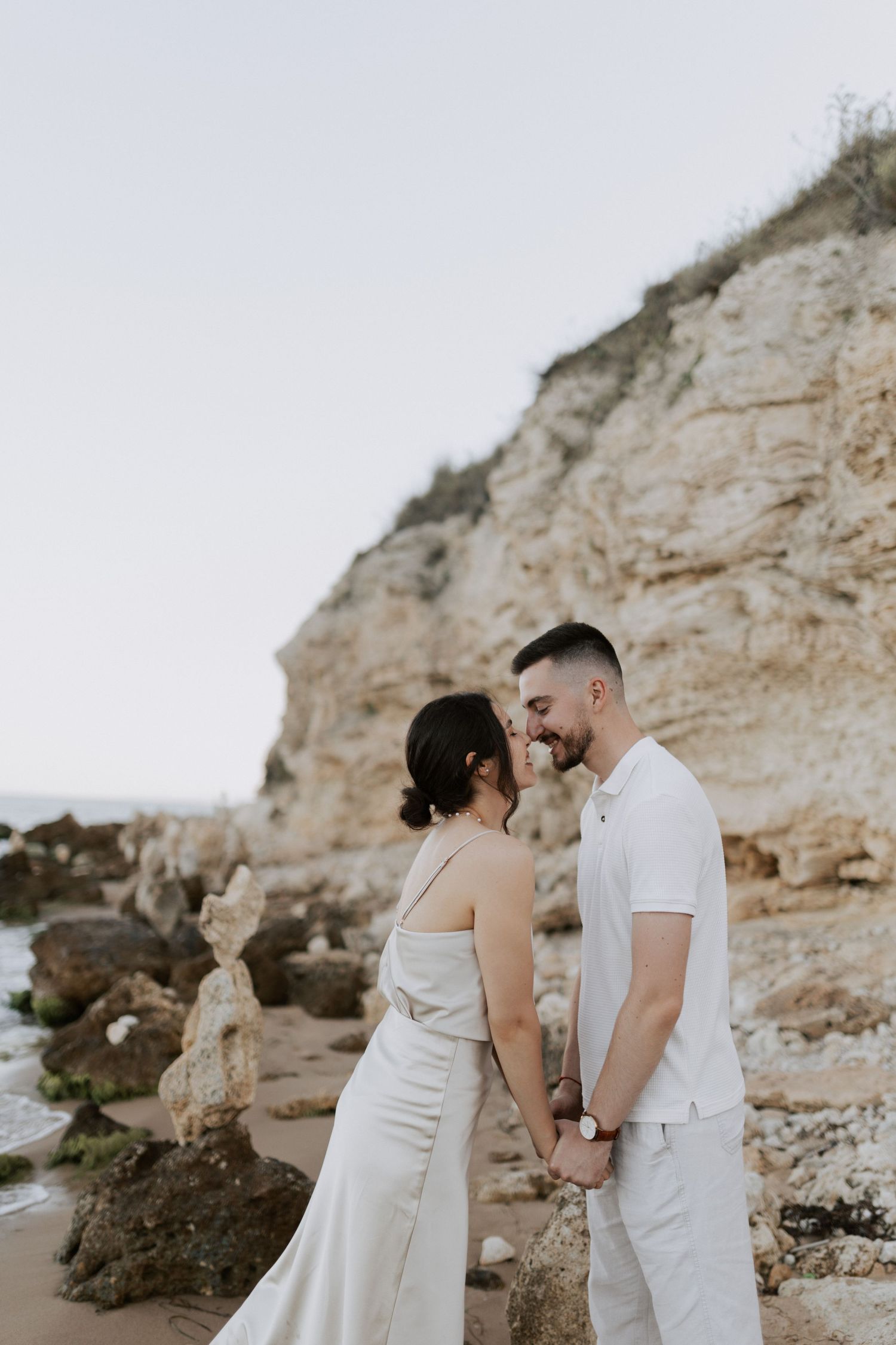 Intimate moment between a couple in white attire against dramatic coastal cliffs.