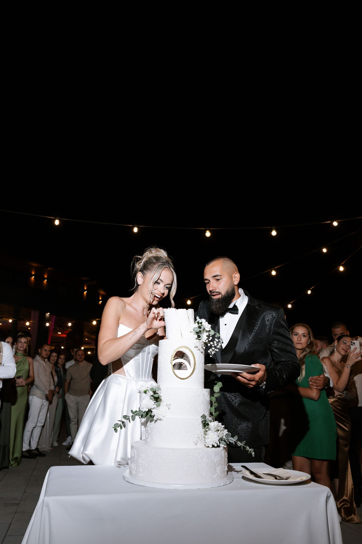Newlywed couple cutting their elegant white wedding cake together at an evening reception with string lights overhead.