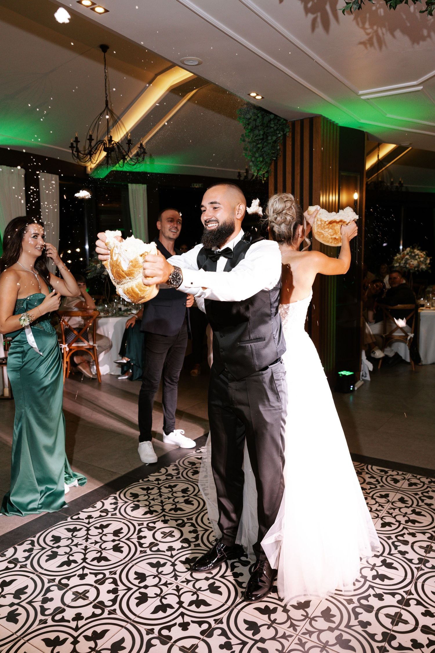 Wedding guests dance and celebrate on a black and white patterned floor at an elegant reception with green accent lighting.