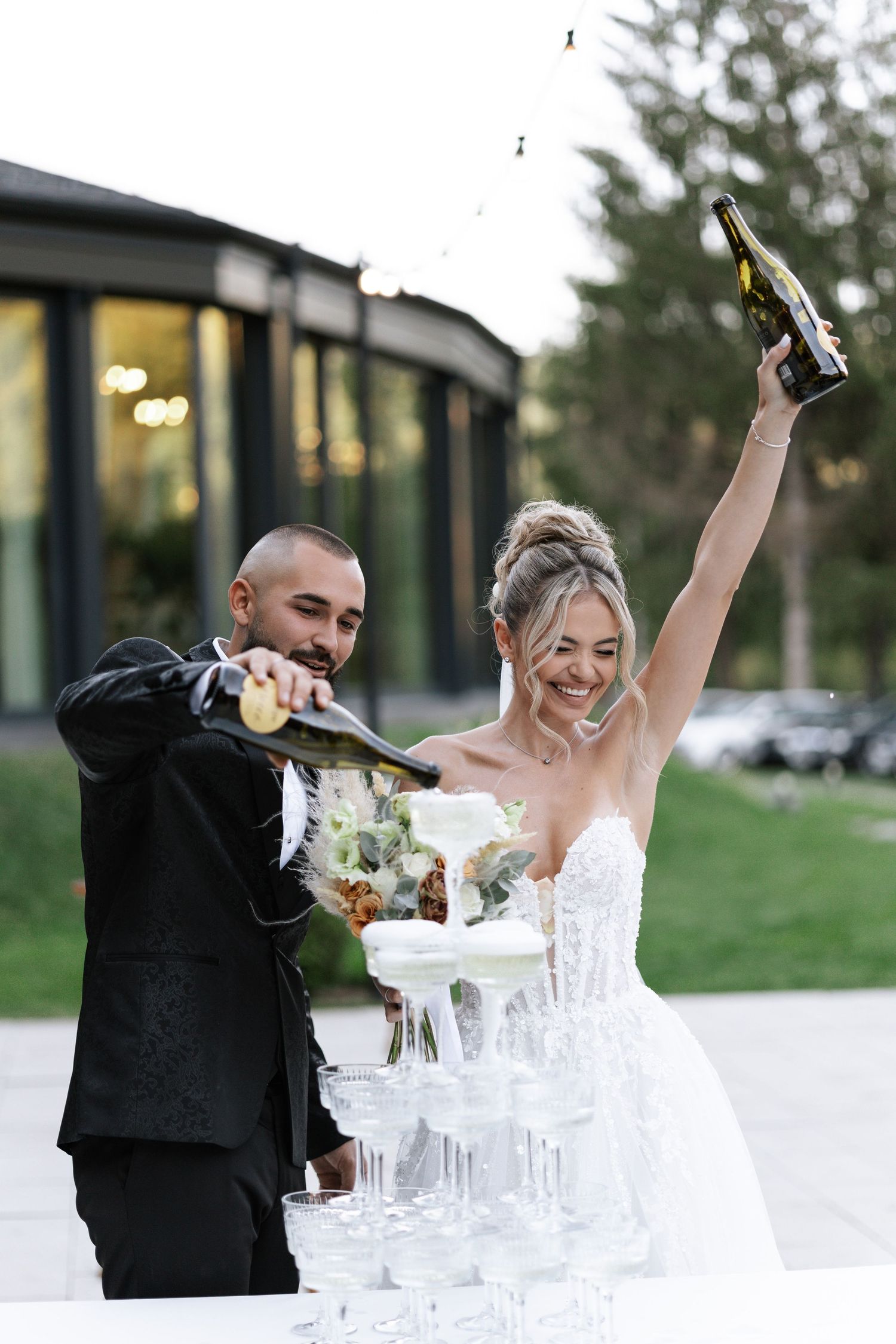 A couple pours champagne into a tower of glasses during their wedding celebration outdoors.