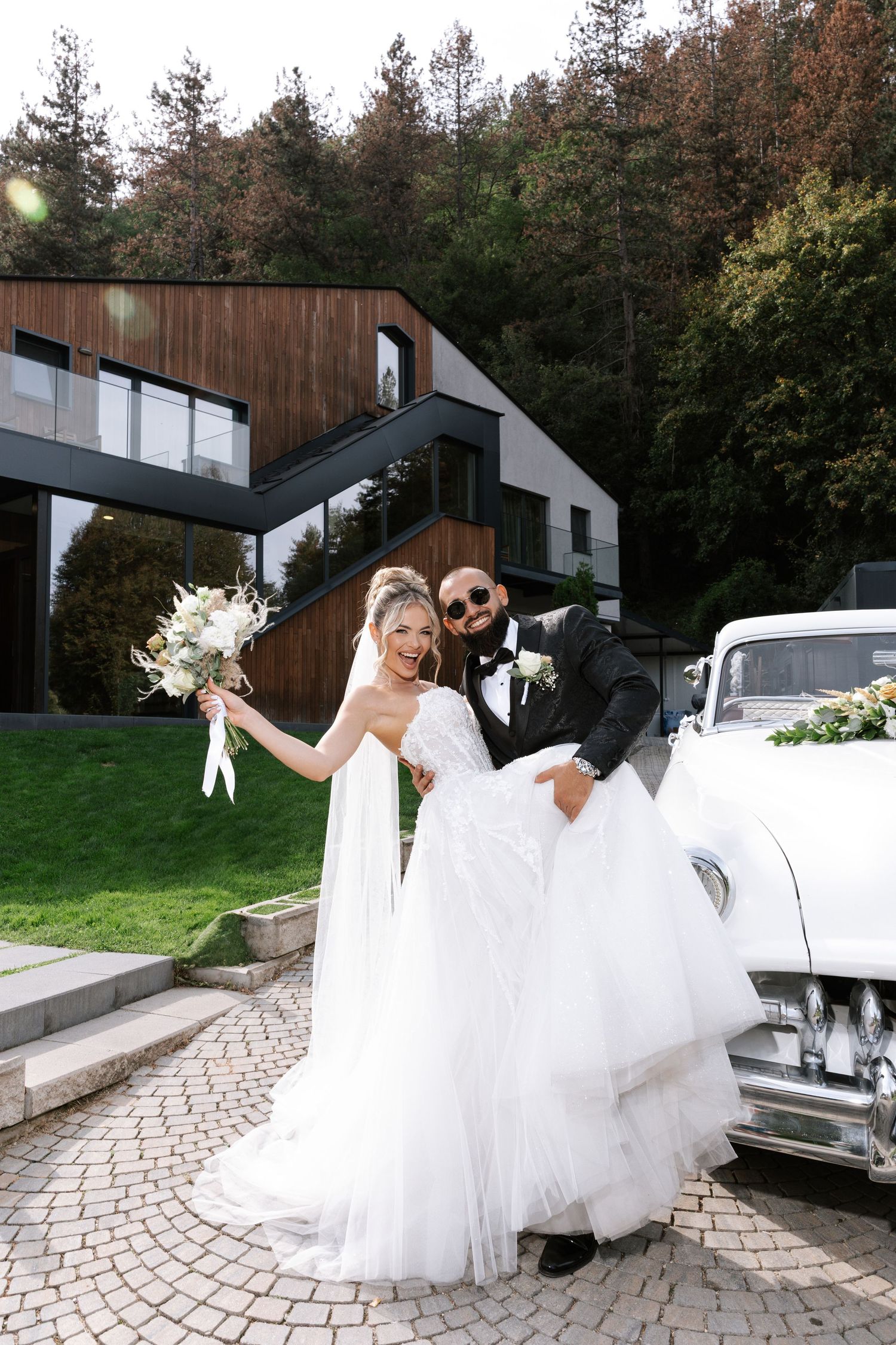 A romantic wedding couple poses by a vintage car on a curved stone driveway surrounded by autumn trees.