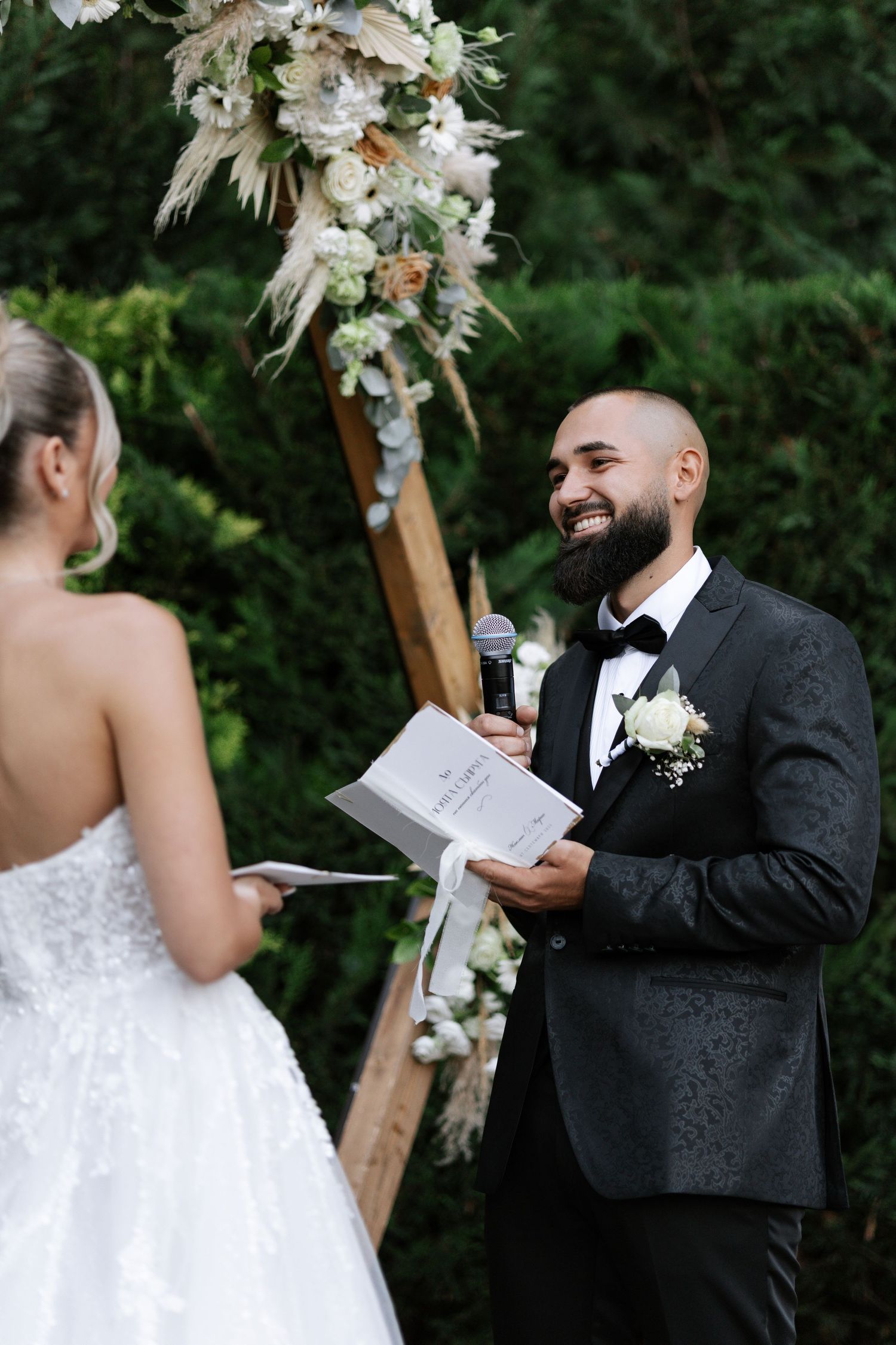 A couple exchanges vows during an outdoor wedding ceremony under a wooden arch decorated with white flowers.