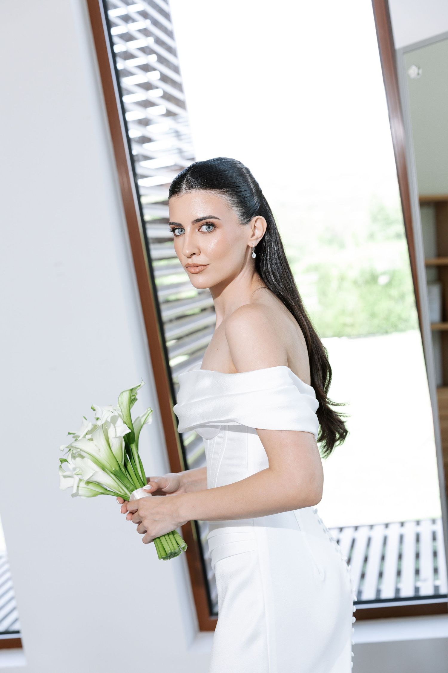 Elegant bride in off-shoulder white dress holds a classic calla lily bouquet while standing by a window.
