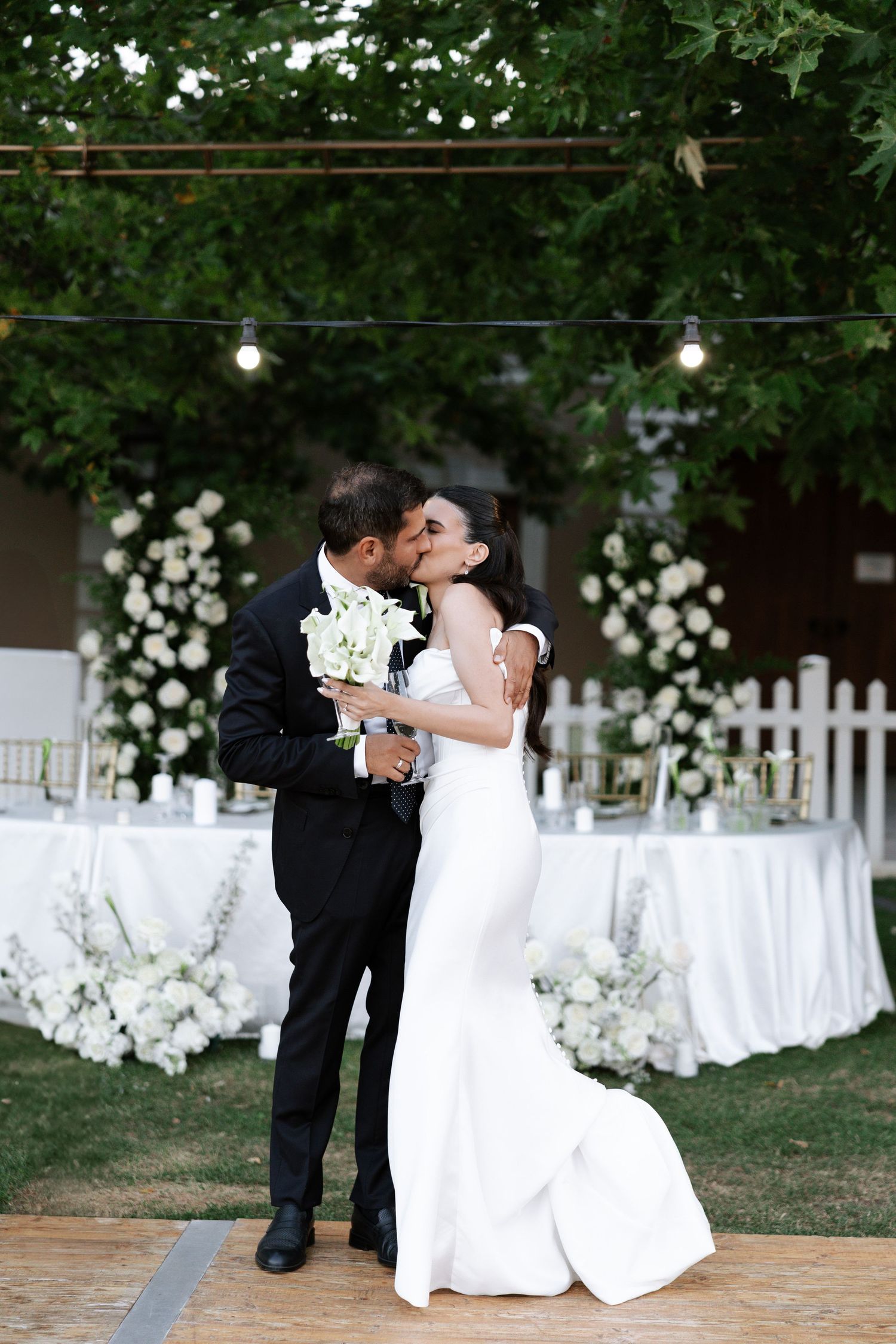 A romantic wedding moment captured on a brick patio with white floral decorations and string lights in the background.