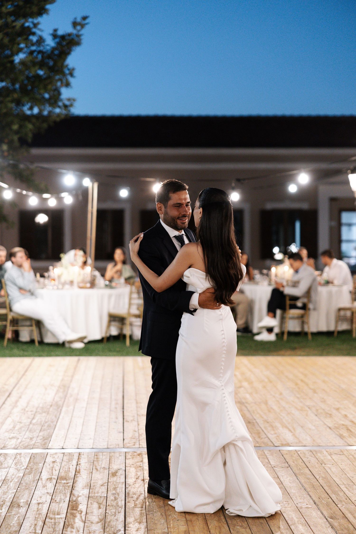 Couple shares romantic moments on wooden deck at outdoor evening wedding reception with string lights overhead.
