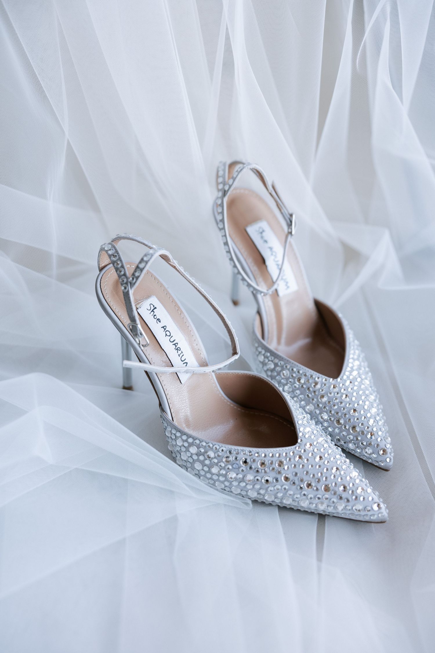 Sparkly silver wedding heels with crystal embellishments placed on white tulle fabric.