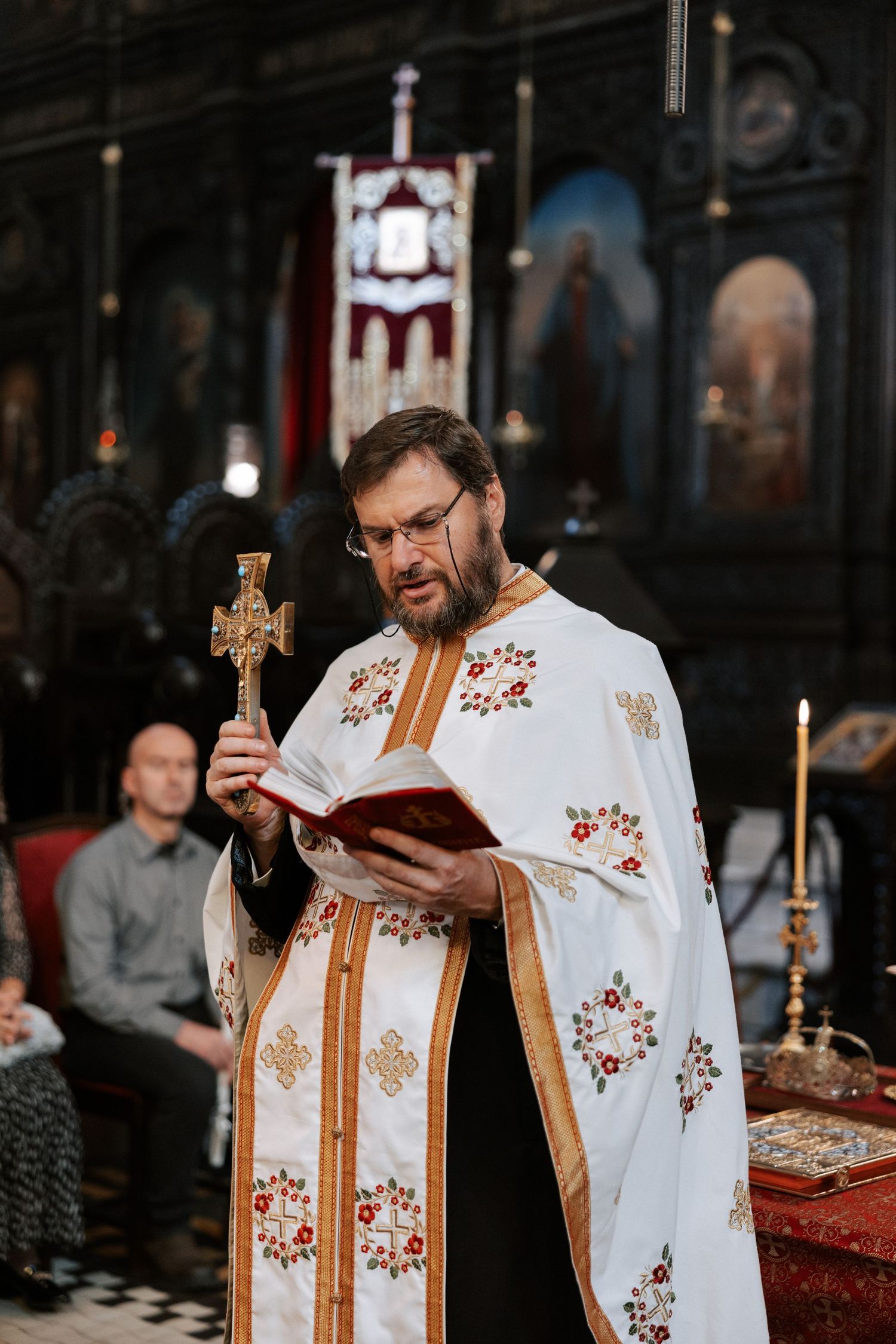 Orthodox priest in white embroidered vestments holds cross and prayer book during church service.