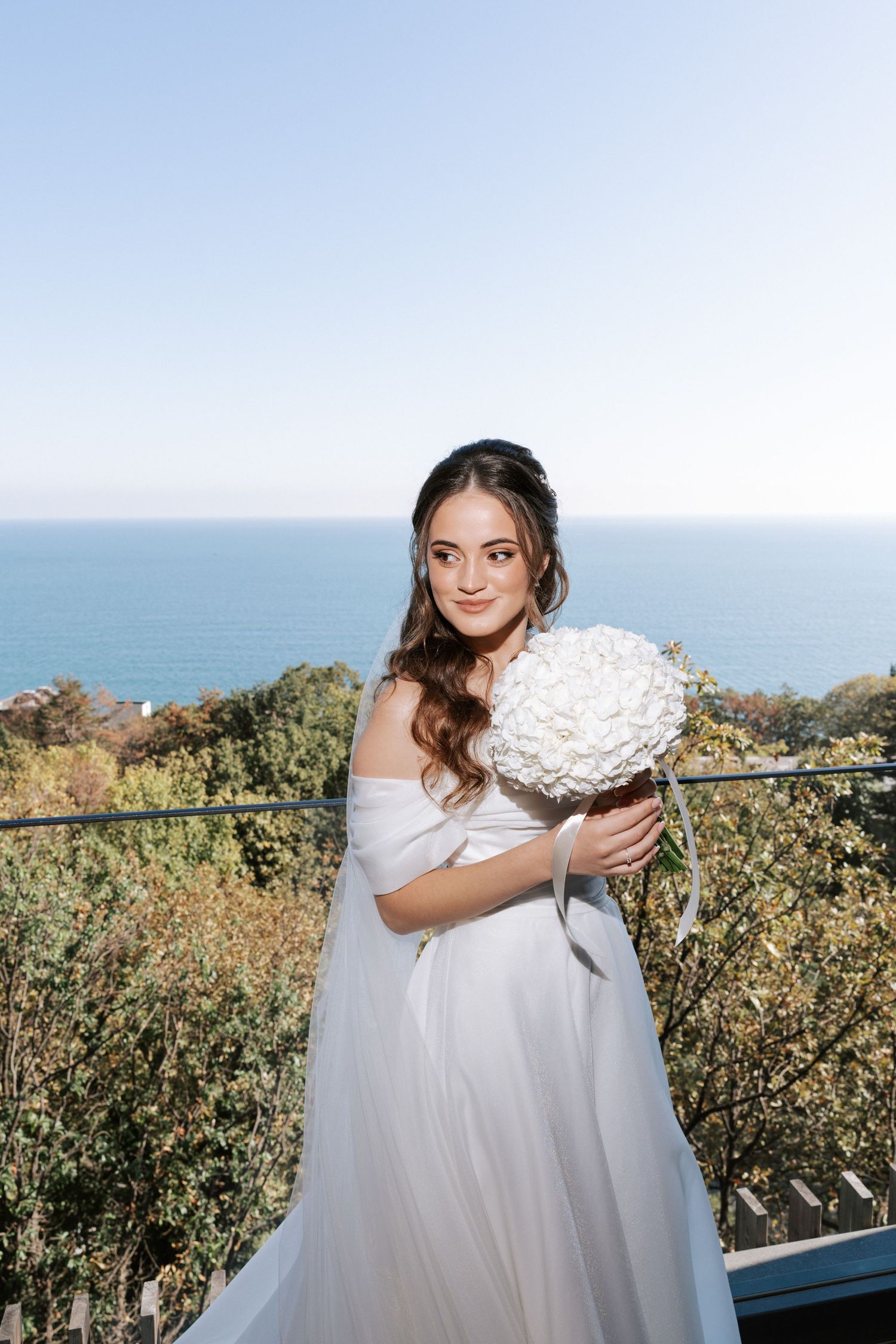 Bride in white dress holding white bouquet against coastal ocean backdrop.