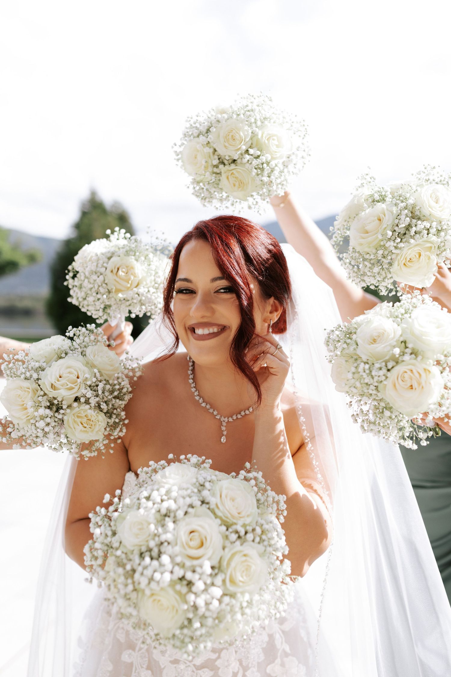 Bride holding white rose and baby's breath bouquets against a lakeside backdrop.