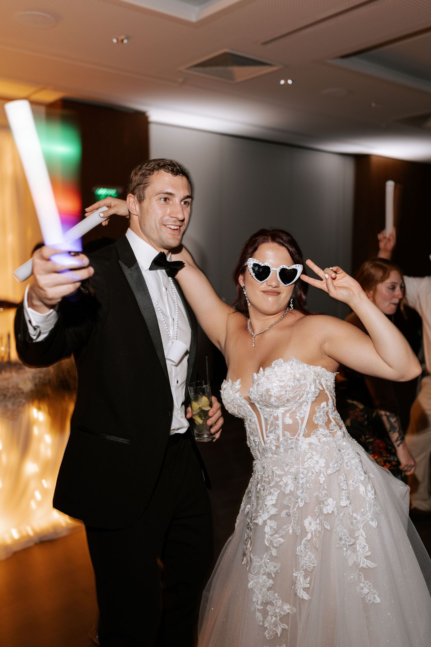 Couple celebrating at wedding reception holding colorful LED glow sticks and wearing sunglasses.