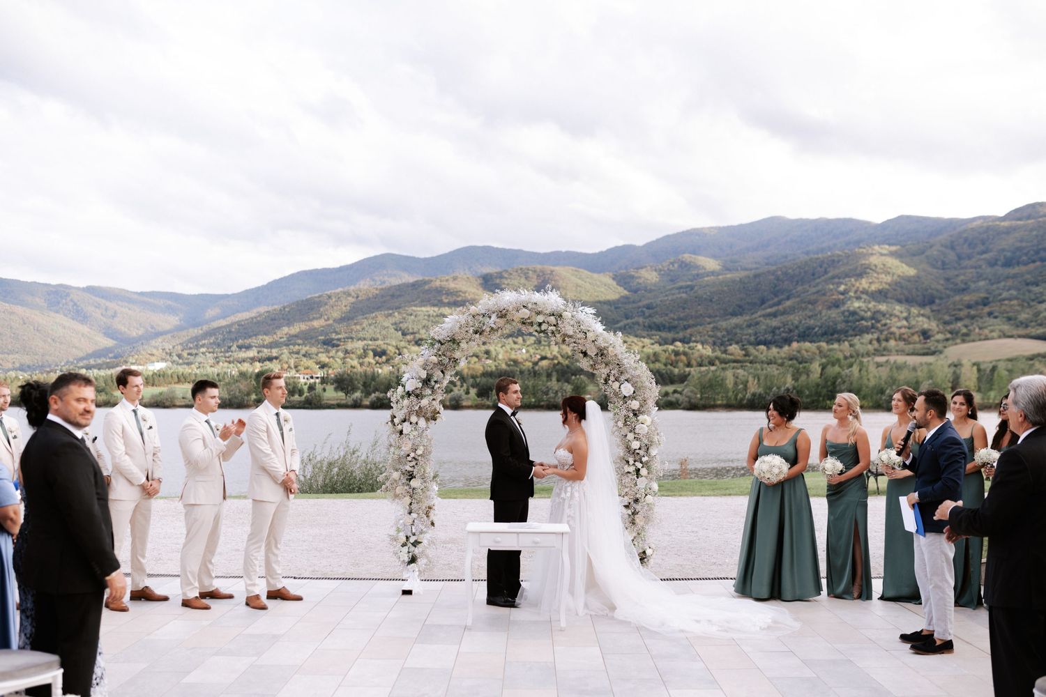 Wedding party in white and sage green attire gathers around floral arch for mountain ceremony.Wedding photographer Bulgaria