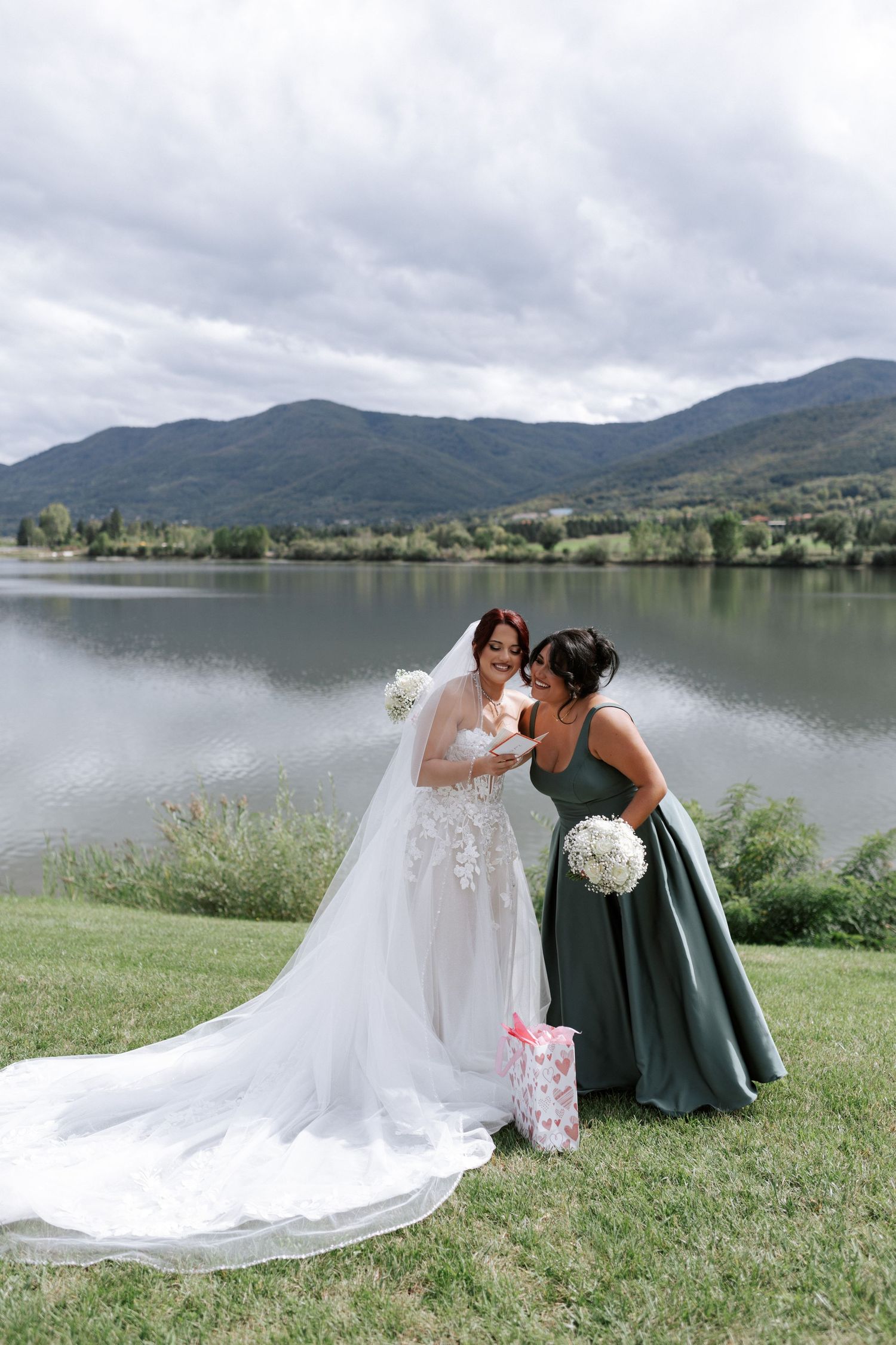 Bride and bridesmaid share laughter while looking at gifts by a scenic mountain lake.