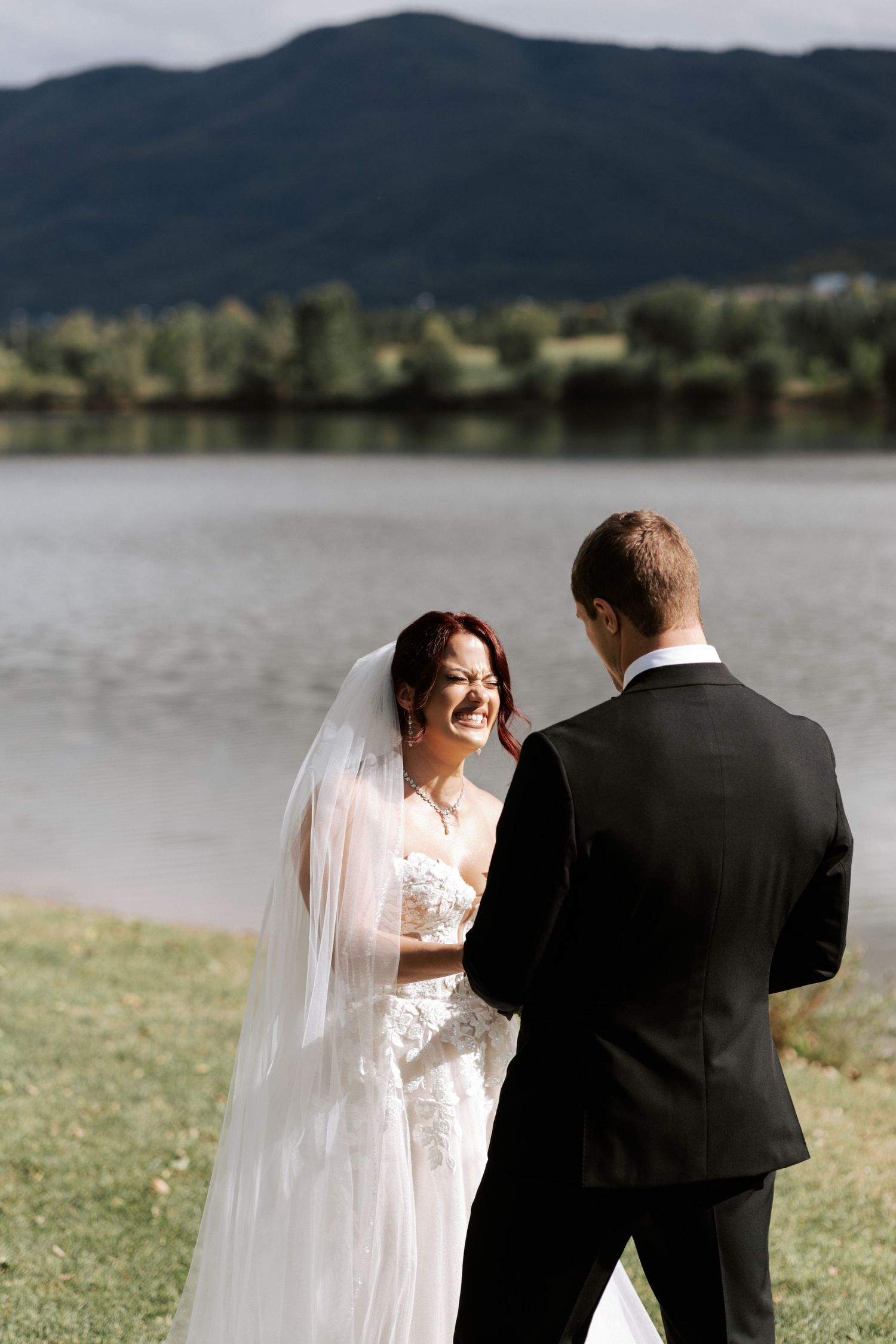 A wedding couple shares a joyful moment by a serene lake with mountains in the background.