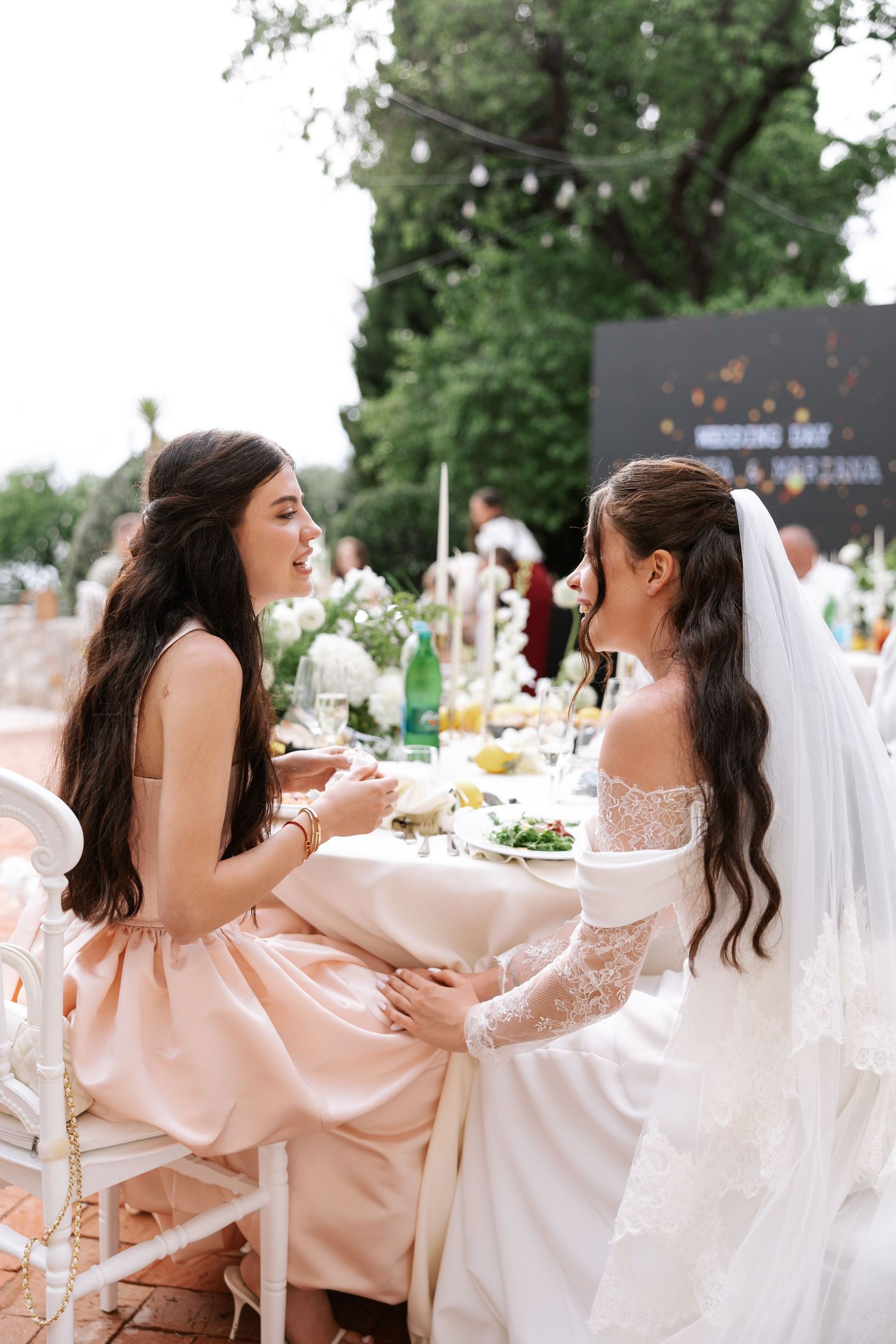 A bride and her bridesmaids in blush pink dresses share joyful moments during an outdoor wedding reception.
