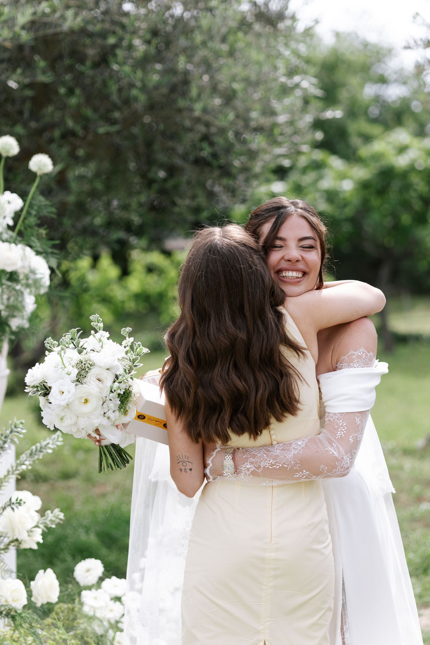 Two people in white dresses embrace warmly in an outdoor garden setting with green foliage in the background.